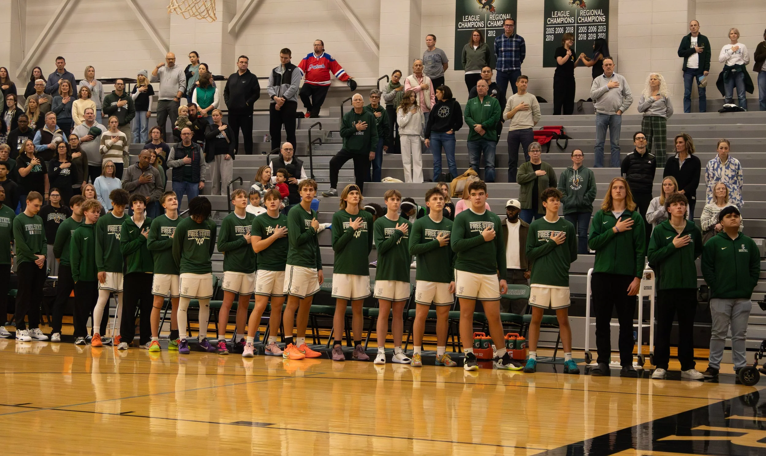 Basketball players in green jerseys standing with their hands over their hearts in a gymnasium during the national anthem, with spectators in the stands behind them.
