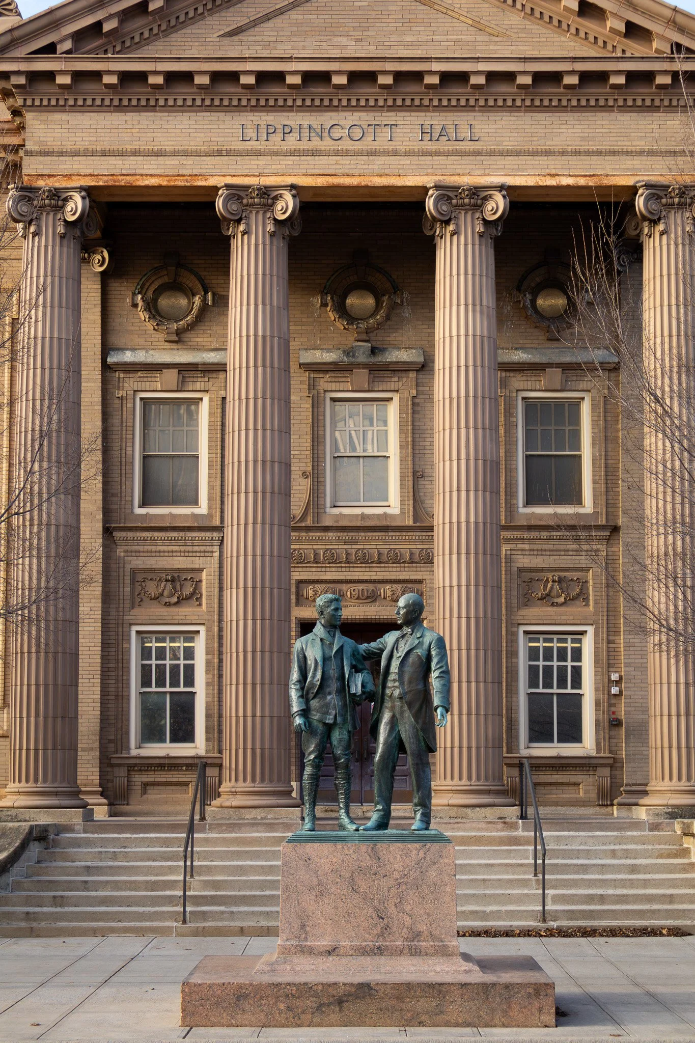 Statue of two men shaking hands outside Lippincott Hall, a historic brick building with large columns and the name sign above the entrance.