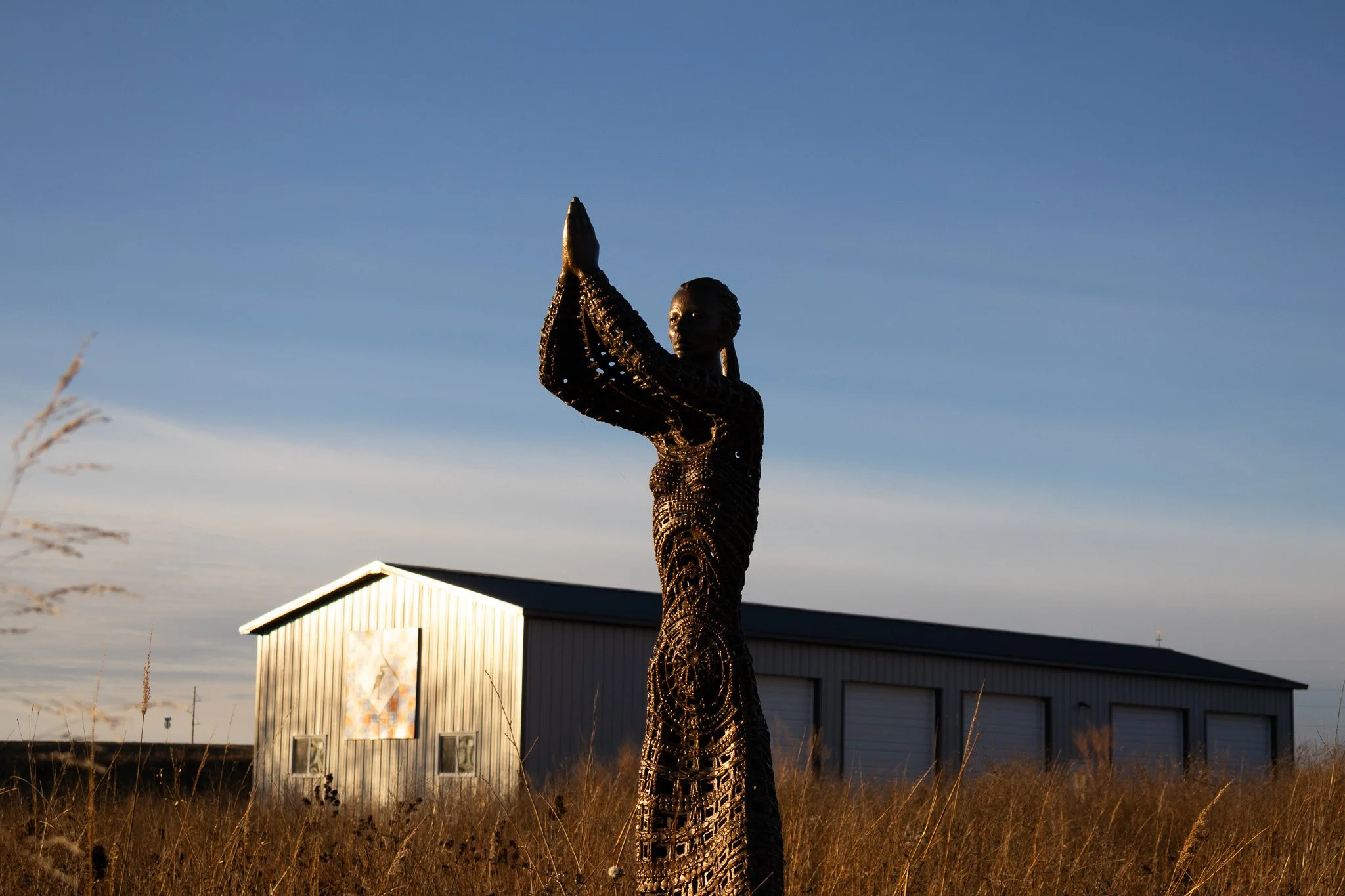 Metal sculpture of a woman with hands pressed together in prayer, standing in tall grass during sunset in front of a metal building with a mural.