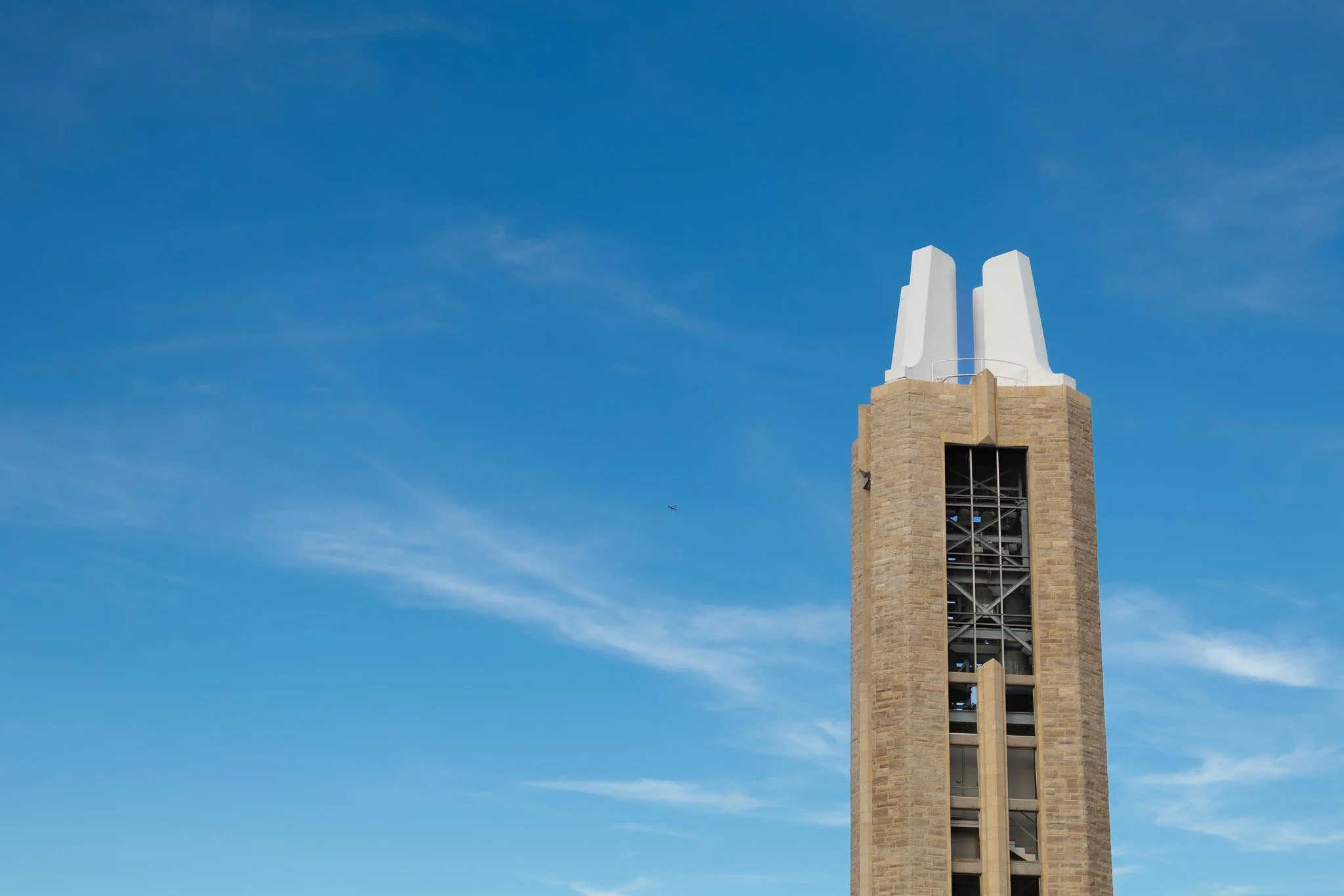 A tall clock tower made of brown brick with white architectural elements at the top, set against a blue sky with wispy clouds.