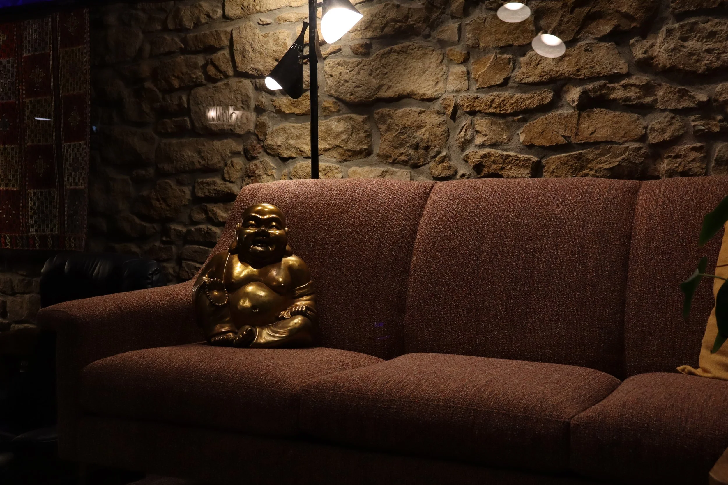 A brown couch with a golden laughing Buddha statue on it, set against a stone wall with a lamp above.