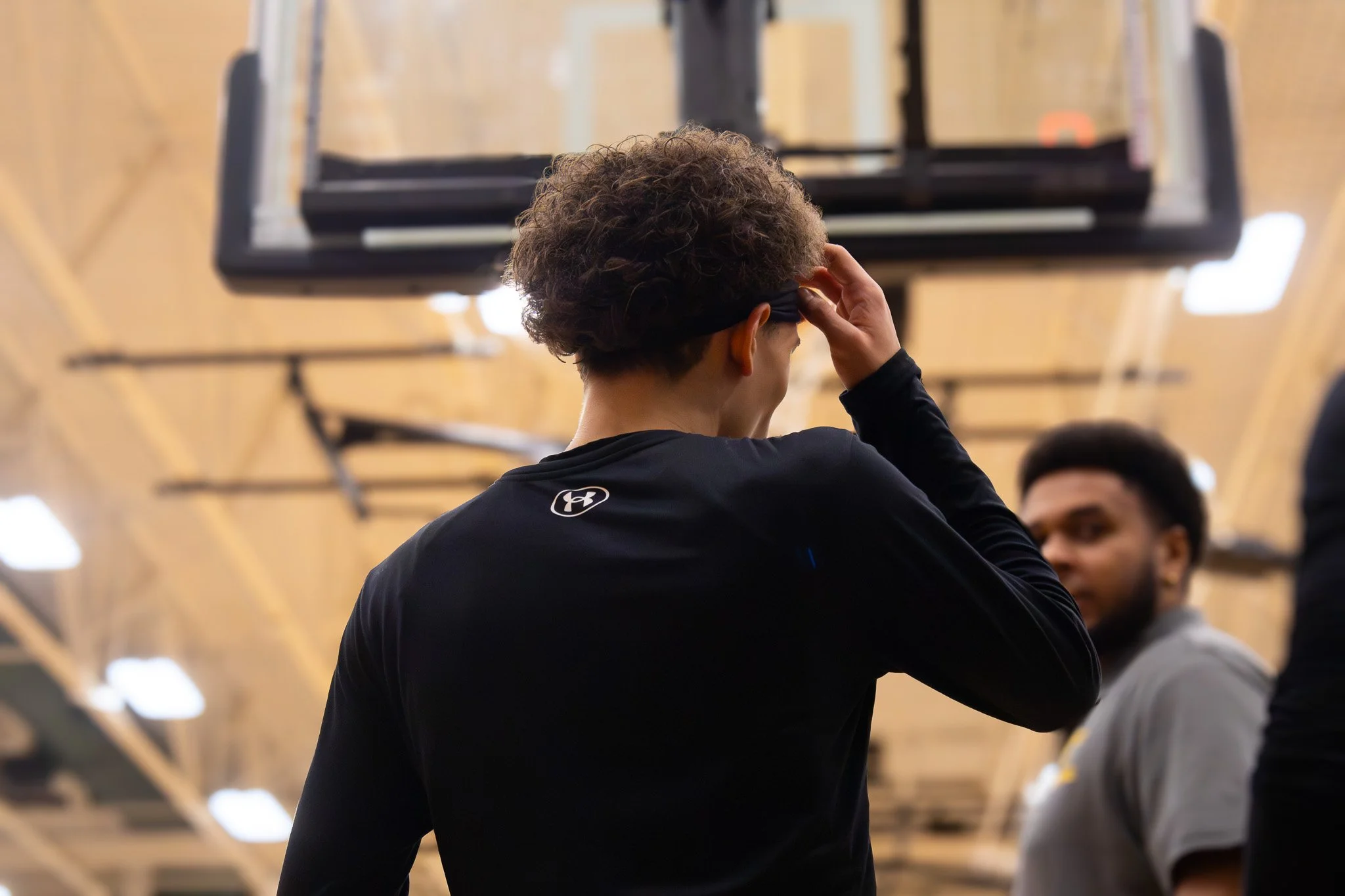 A young man with curly hair wearing a black long sleeve shirt with Under Armour logo, standing inside a gym, adjusting his headband while looking towards another person in the background.