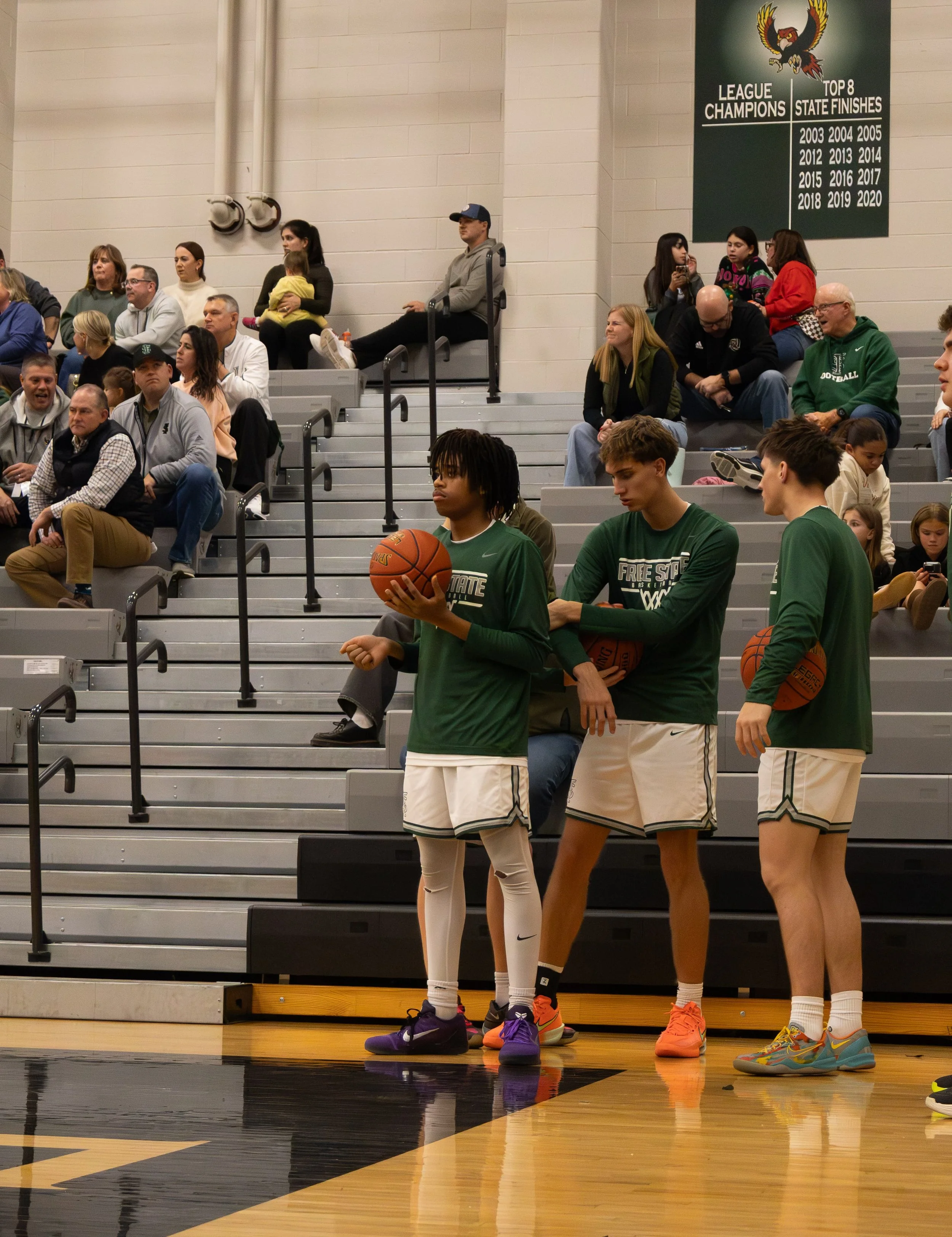 Three basketball players in green uniforms waiting on the court, holding basketballs, while spectators watch from the bleachers in a gymnasium.