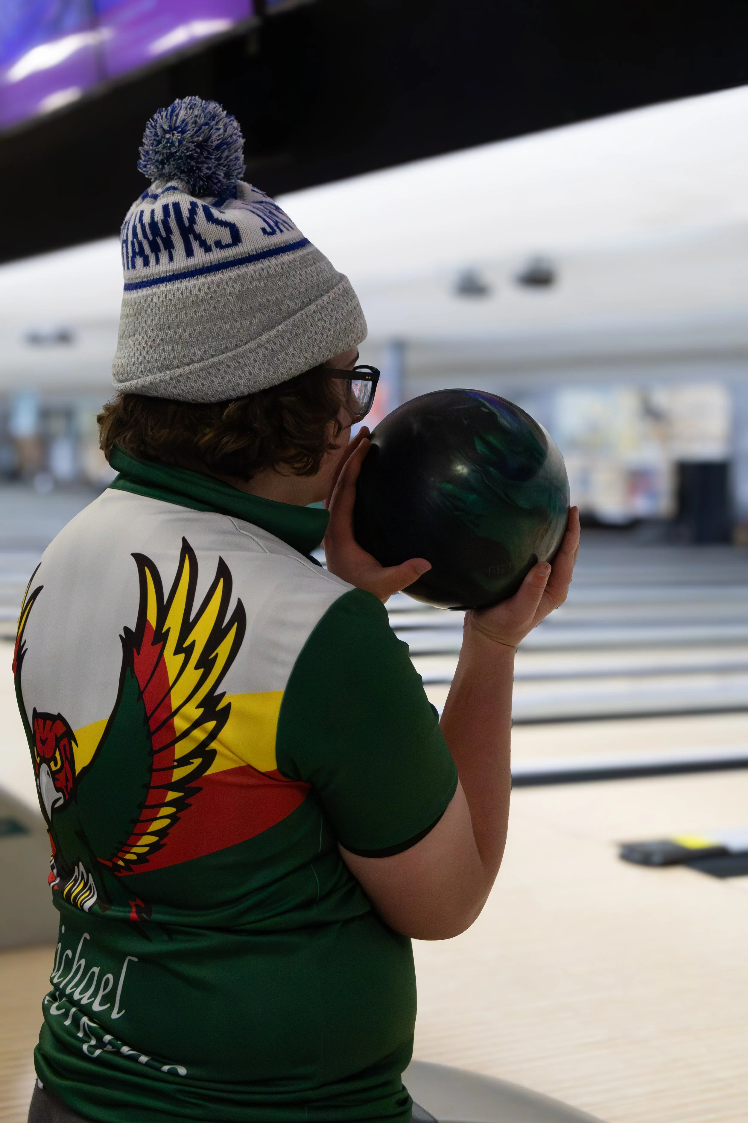 Person holding a bowling ball at a bowling alley, wearing a gray knit hat with a pom-pom and a shirt with a colorful eagle graphic.