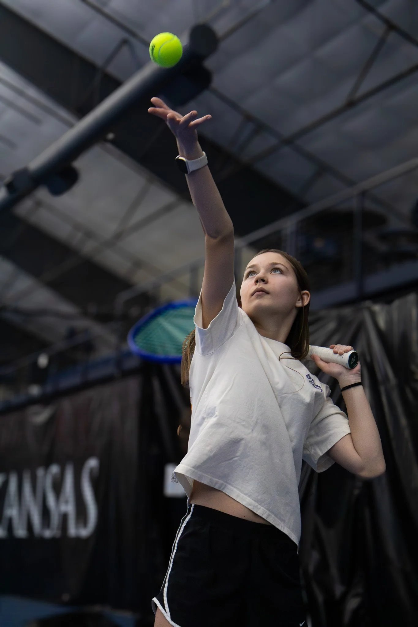 Young girl playing tennis, about to serve, indoors, wearing a white shirt and black shorts, holding a tennis racket, with a green tennis ball in the air.