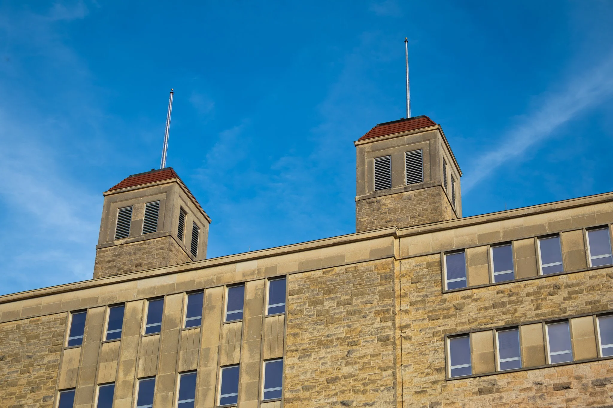 A beige stone building with two square towers with red roofs and black vent windows, set against a bright blue sky with few clouds.