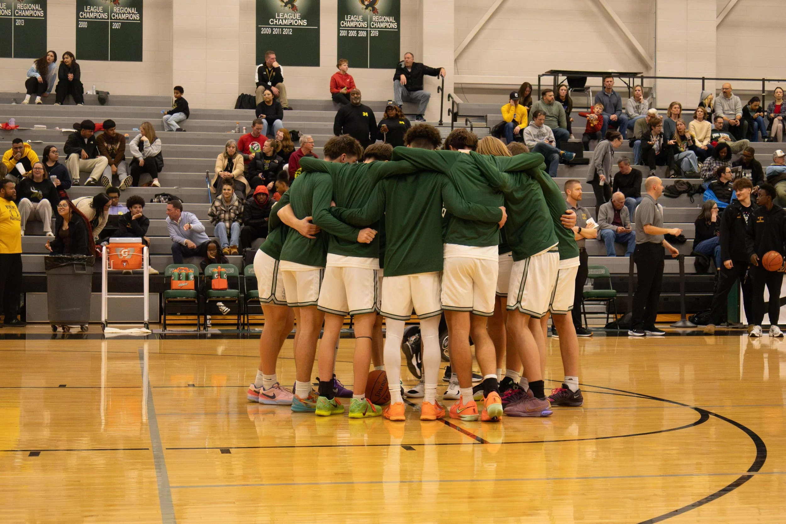 A basketball team in green and white uniforms huddles together on the court during a game. The gym is filled with spectators sitting on bleachers and watching.