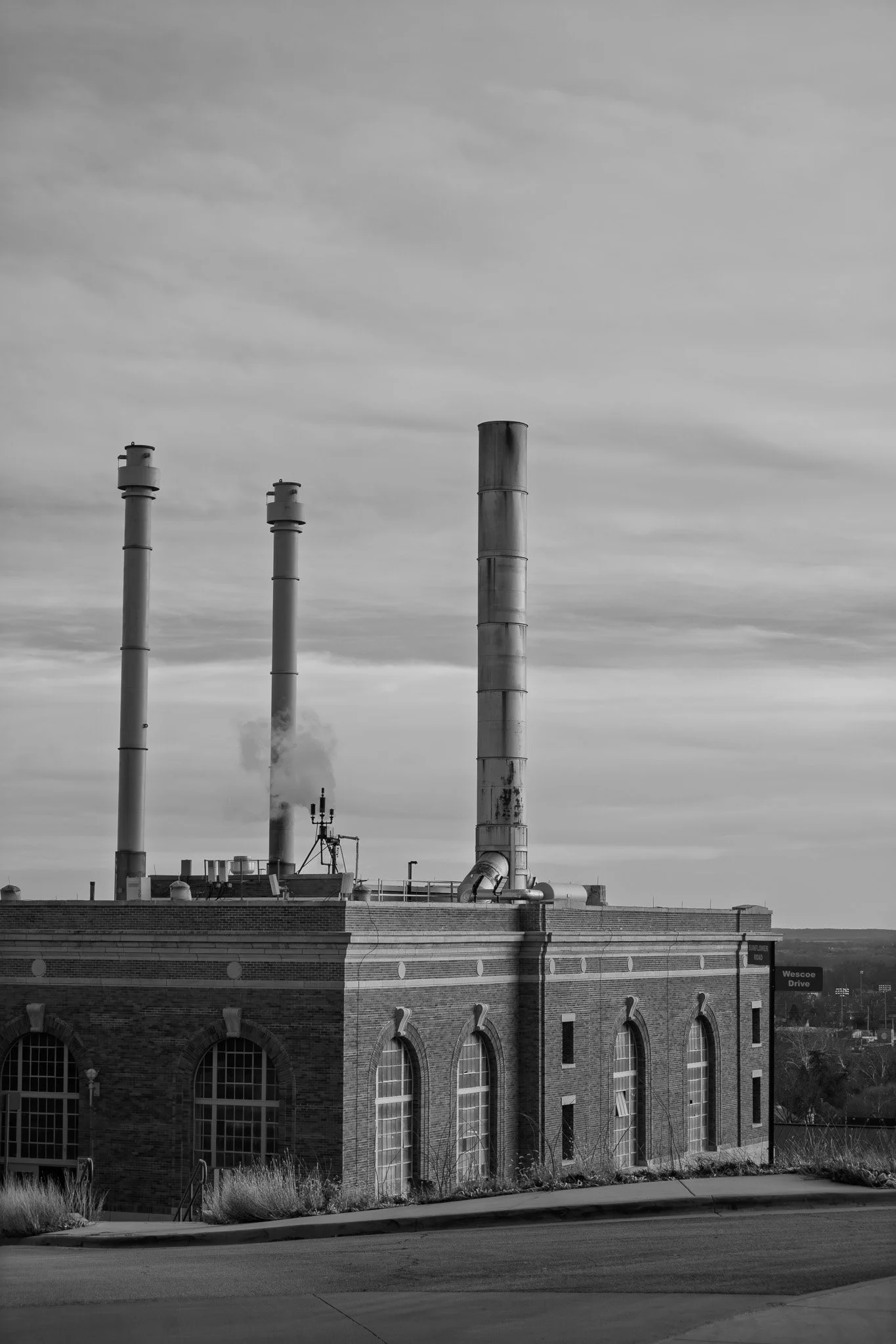 Black and white photo of an industrial building with three tall smokestacks, two emitting smoke, against a cloudy sky.