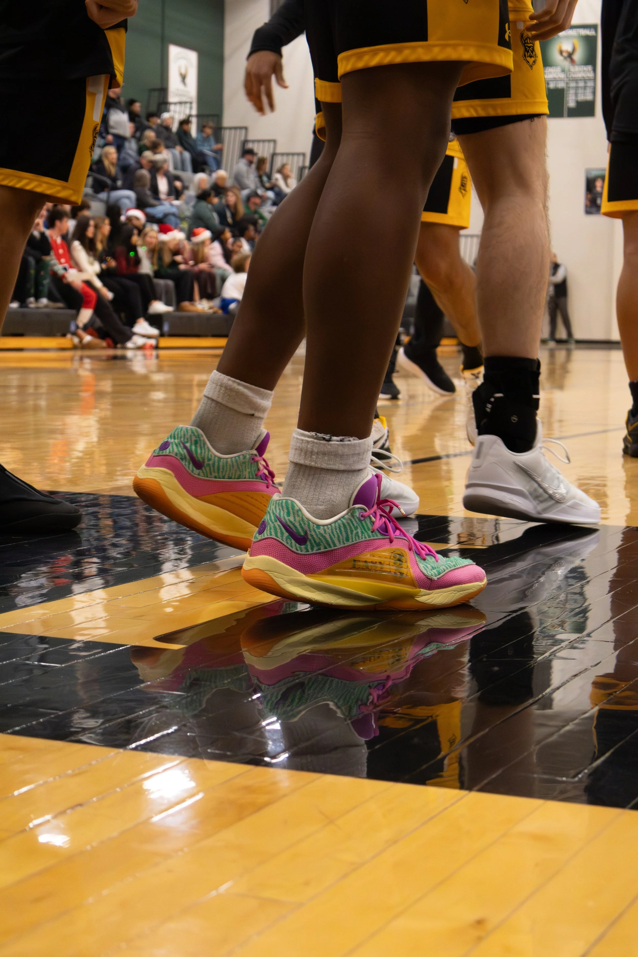 Close-up of a person wearing colorful sneakers, standing on a basketball court during a game with spectators in the background.