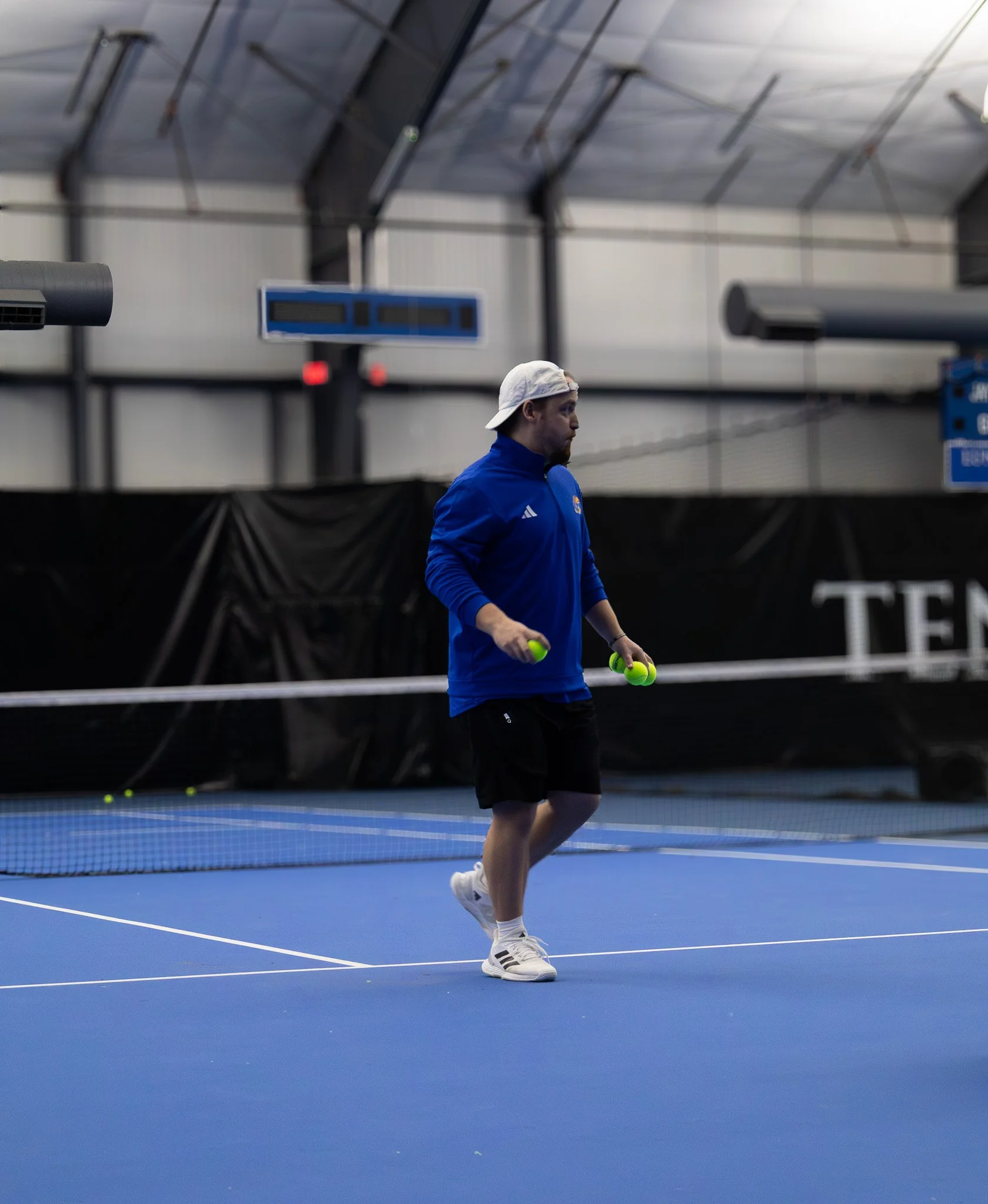 A person in athletic clothing standing on an indoor tennis court holding tennis balls.