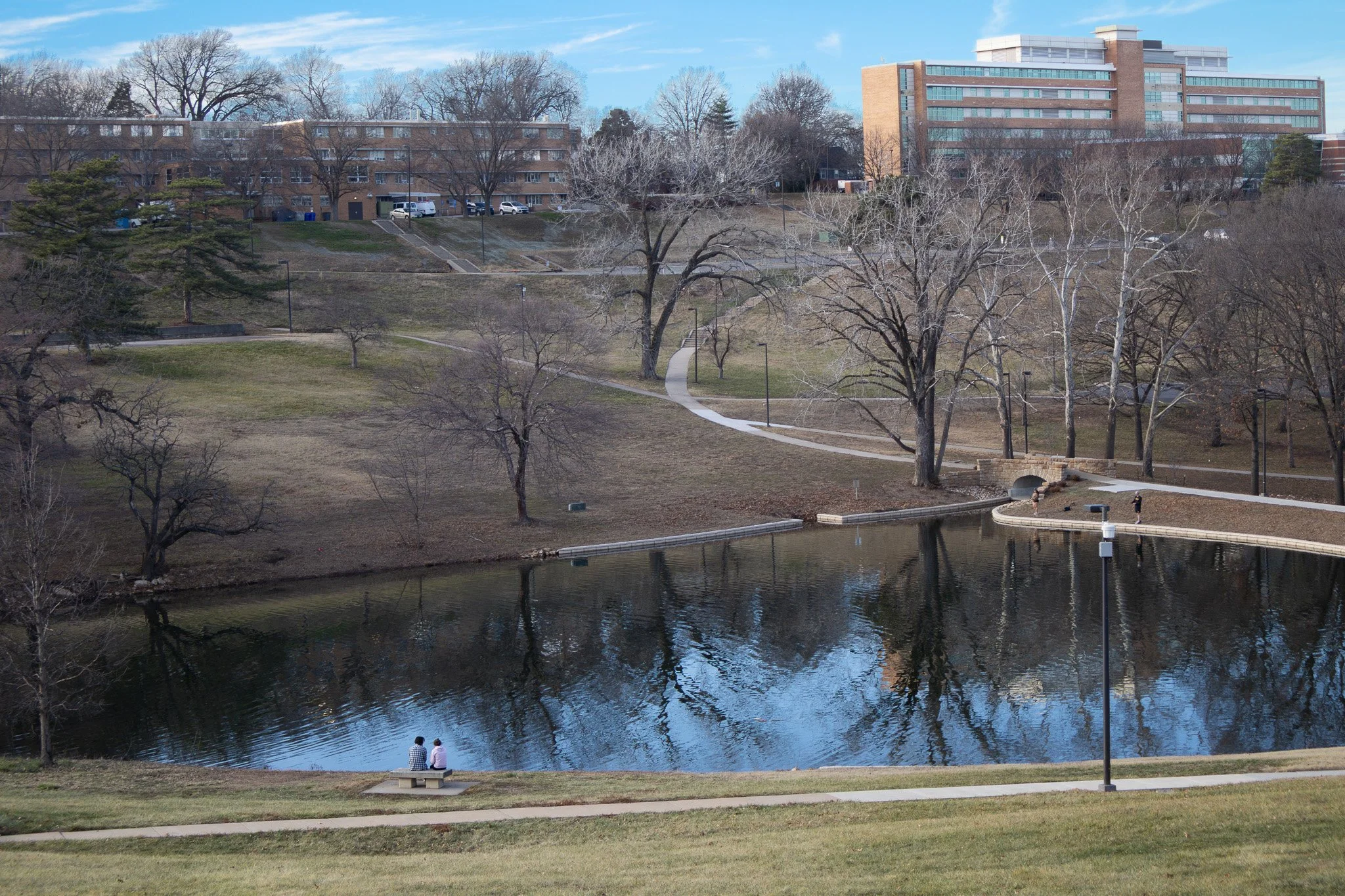 A park with a pond, leafless trees, a paved walking path, and two people sitting on a bench near the water. In the background are modern multi-story buildings and a partly cloudy sky.