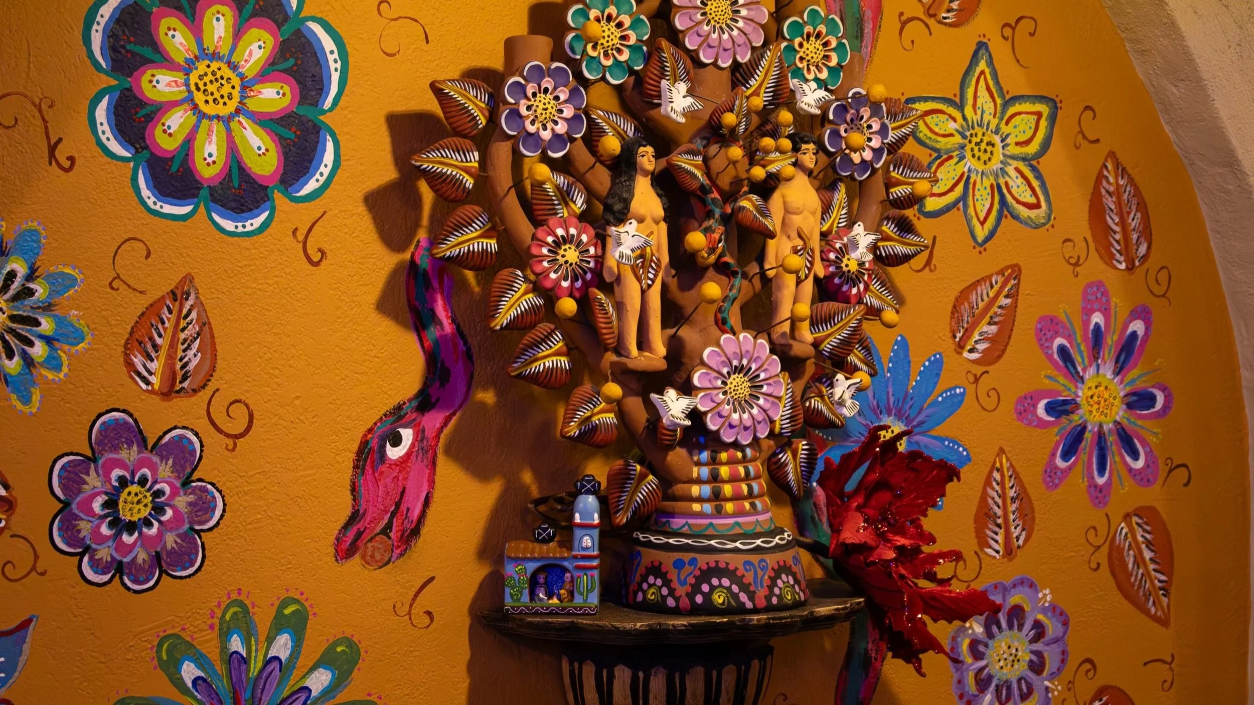 Colorful Mexican folk art decoration on a yellow wall, featuring a large floral arrangement with painted figures, flowers, butterflies, and a pink snake, surrounded by painted floral and leaf motifs.