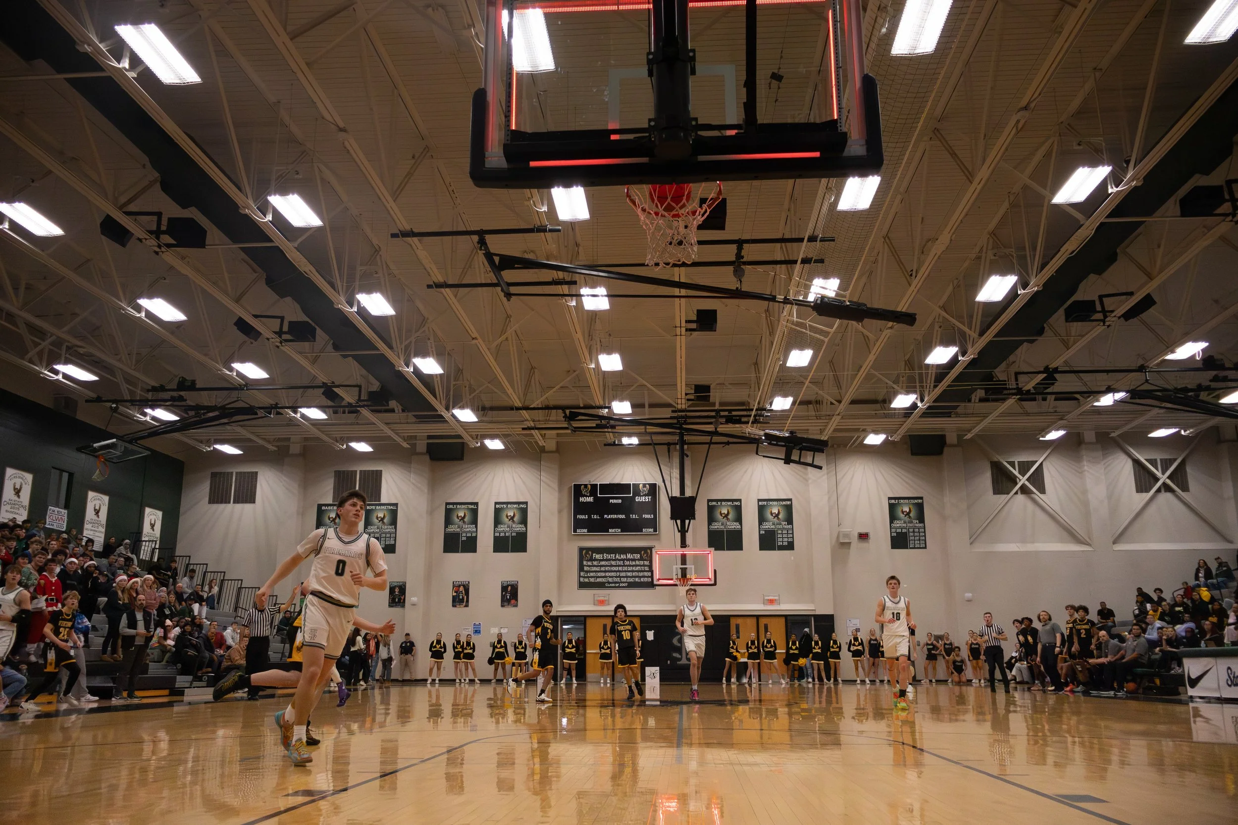 A basketball game is taking place in a gymnasium with players running on the court, spectators seated on the sides, and a scoreboard visible in the background.