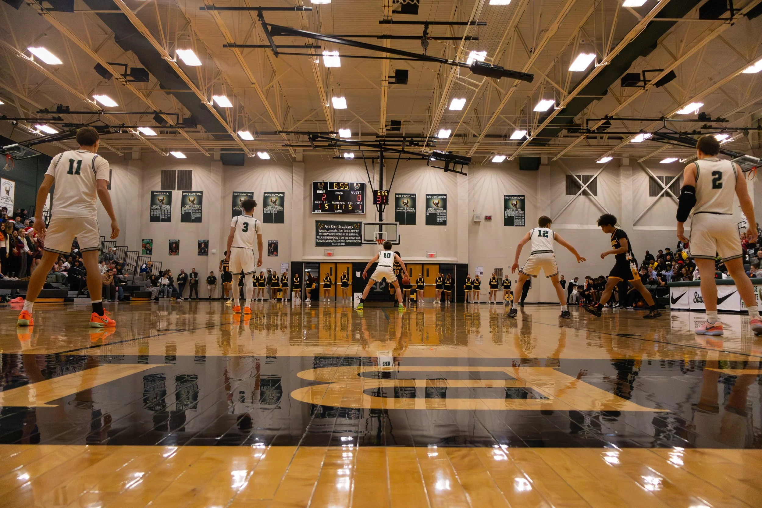 High school basketball game in a gymnasium, players on the court preparing for free throw, scoreboard displaying 6 minutes and 55 seconds remaining in the game, audience seated on bleachers, cheerleaders on the sidelines.