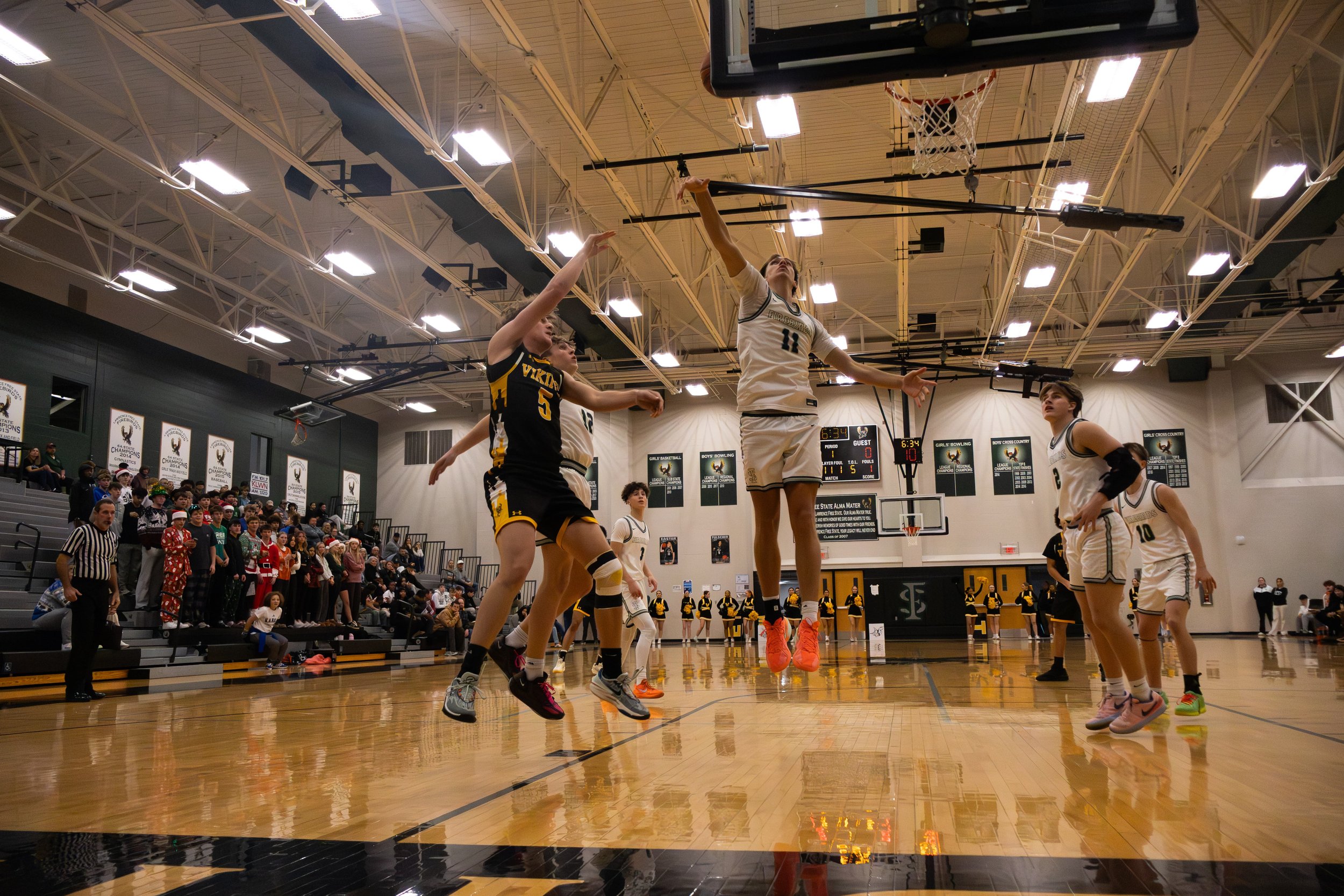 Women's basketball game in gymnasium, players jumping for ball, audience watching, scoreboard showing 6:34 remaining, gym decorated with banners.