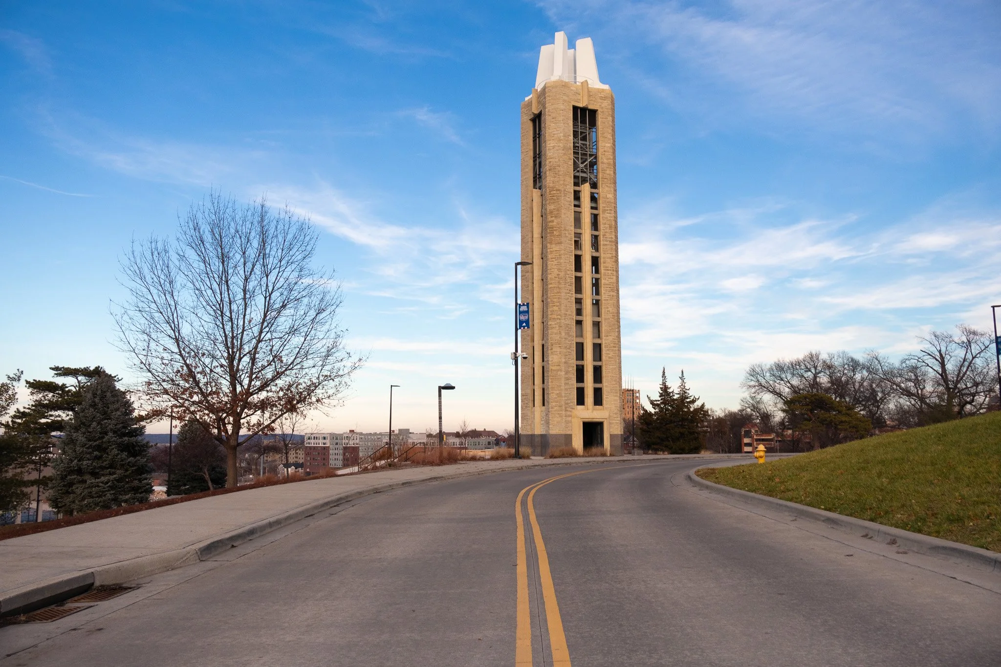 Tall clock tower on a curved road with trees and a clear sky in the background.