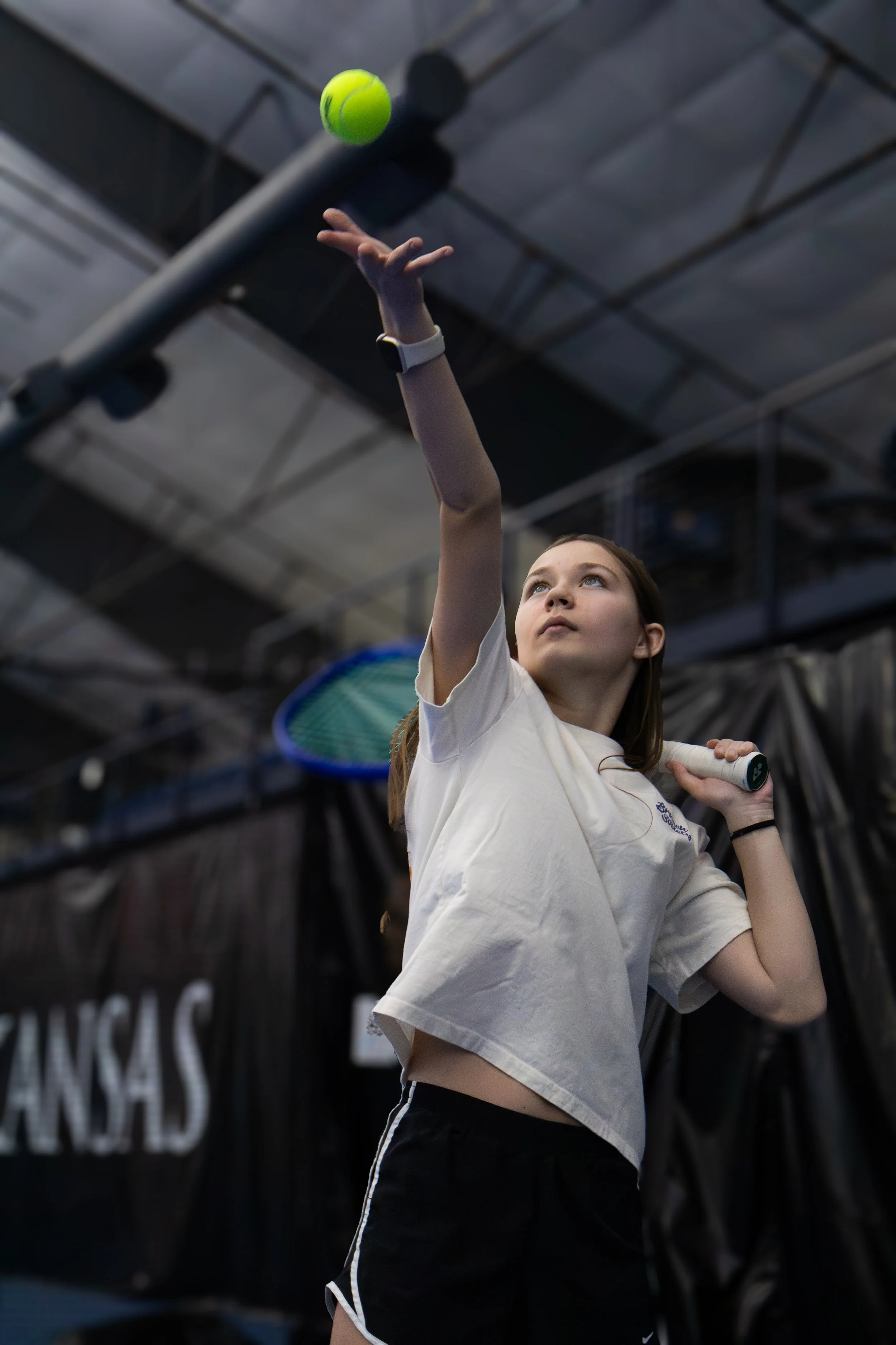 Young girl playing tennis indoors, about to hit a tennis ball with a racket.