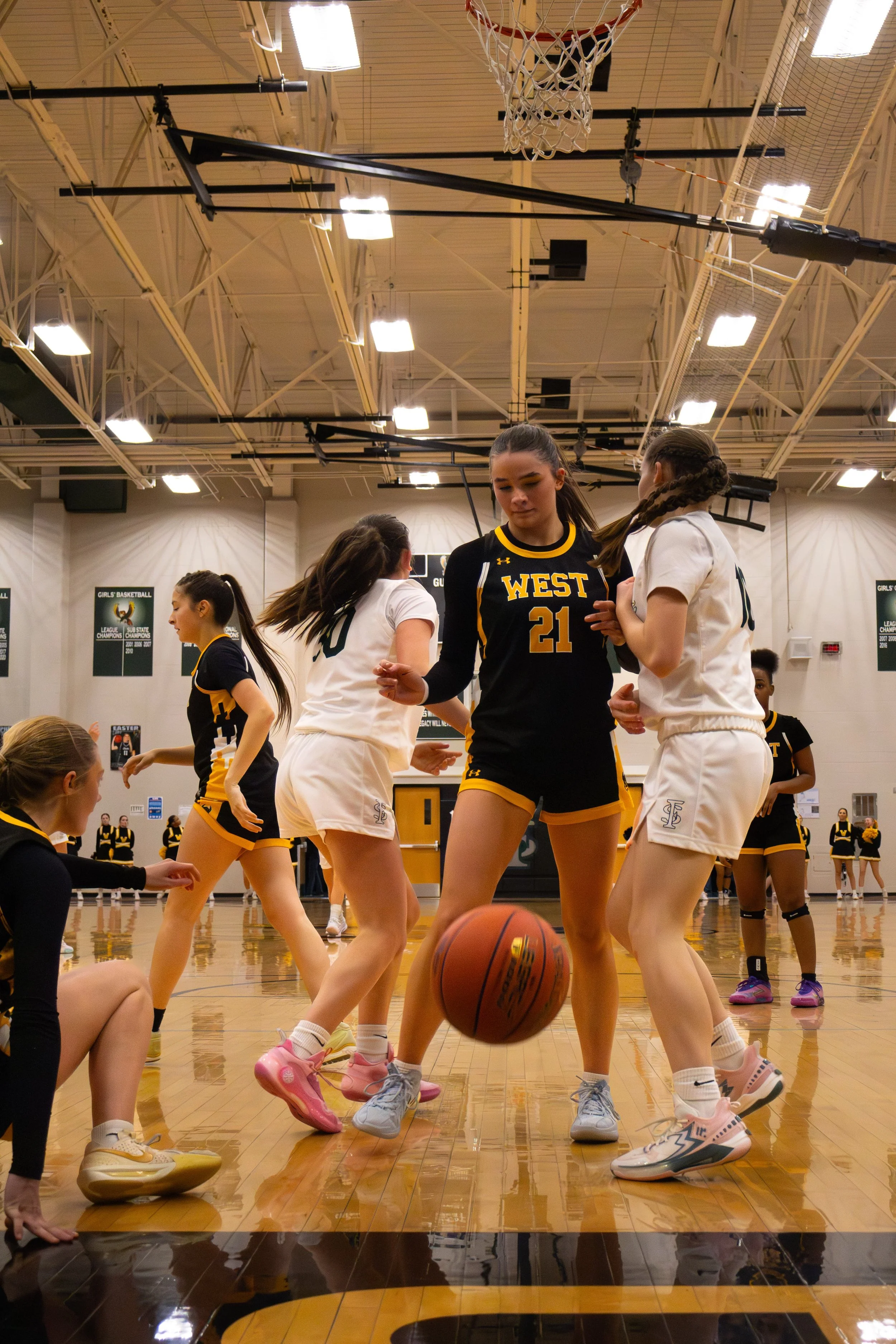 Girls' basketball team during a game or practice in a gymnasium, with one girl in a black jersey holding a basketball, surrounded by teammates in white and black uniforms.