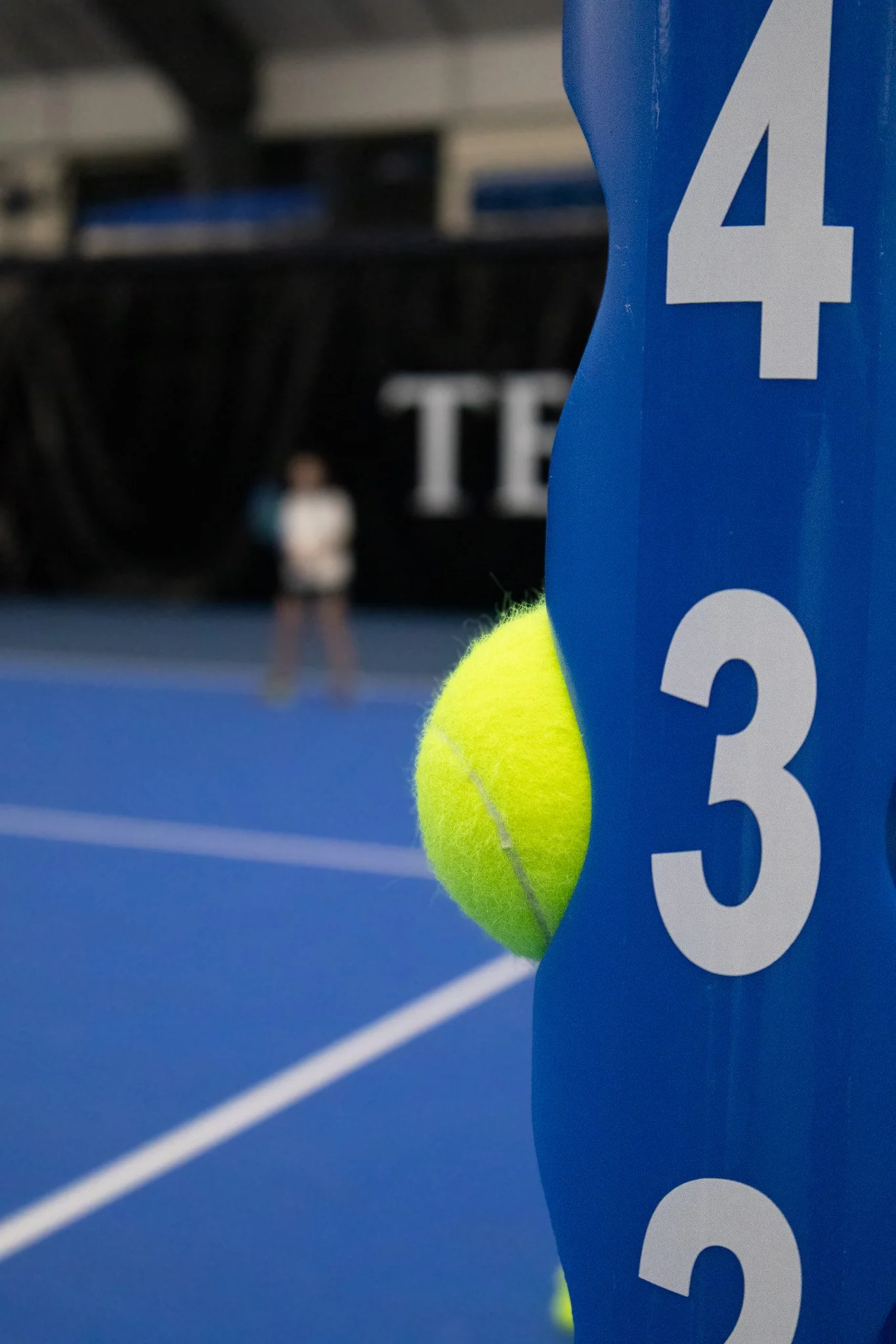 Close-up of a yellow tennis ball partially hidden behind a blue tennis net post with white numbers, on a blue tennis court with a blurred person in the background.