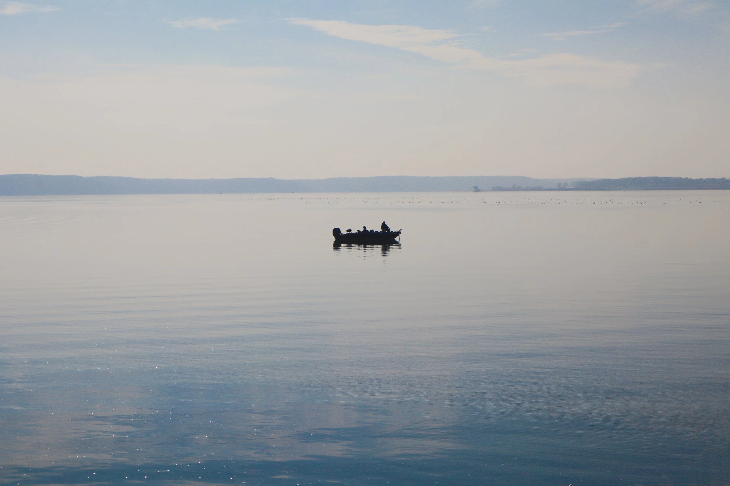 A small boat with three people on it floating on a calm, expansive body of water during daytime with a distant shoreline and some clouds in the sky