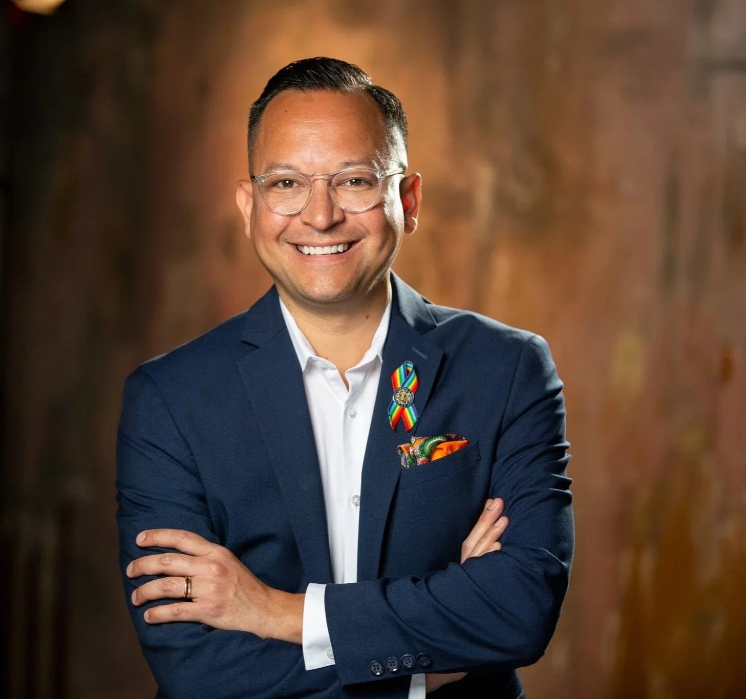 A smiling man with glasses in a navy suit and white shirt, standing with arms crossed, wearing a rainbow ribbon pin and a colorful pocket square, against a blurred warm-toned background.