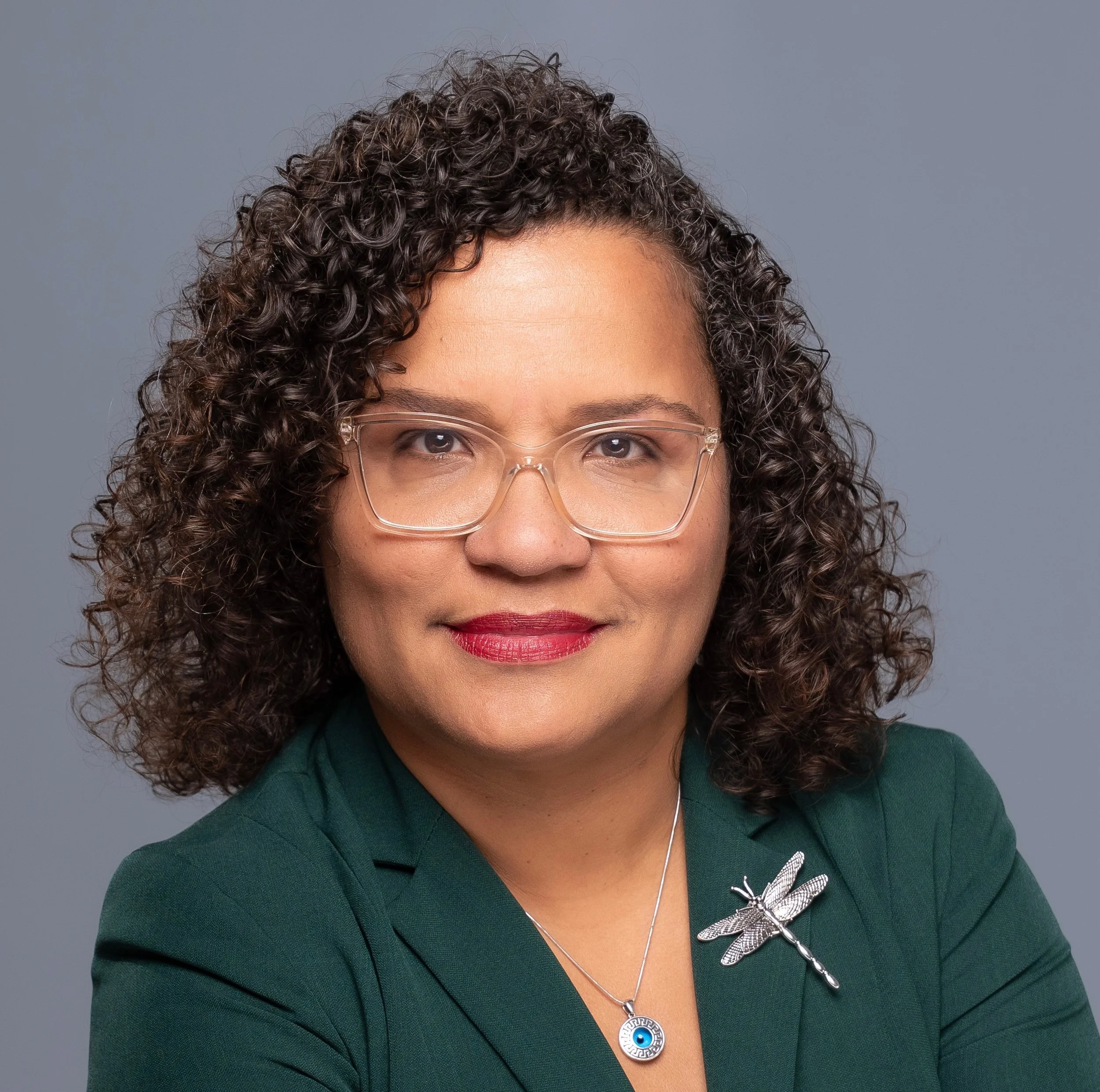 A woman with curly brown hair, glasses, and red lipstick, wearing a green blazer with a dragonfly brooch and a necklace with an eye charm, posing against a neutral background.