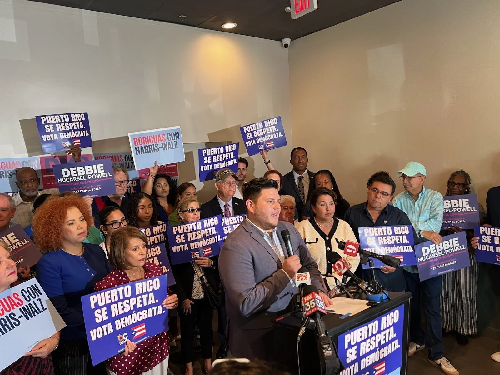 A man in a suit speaking at a press conference with microphones, surrounded by people holding signs that say "Puerto Rico se respeta. Vota demócrata" and "Debbie Mucarsel-Powell."