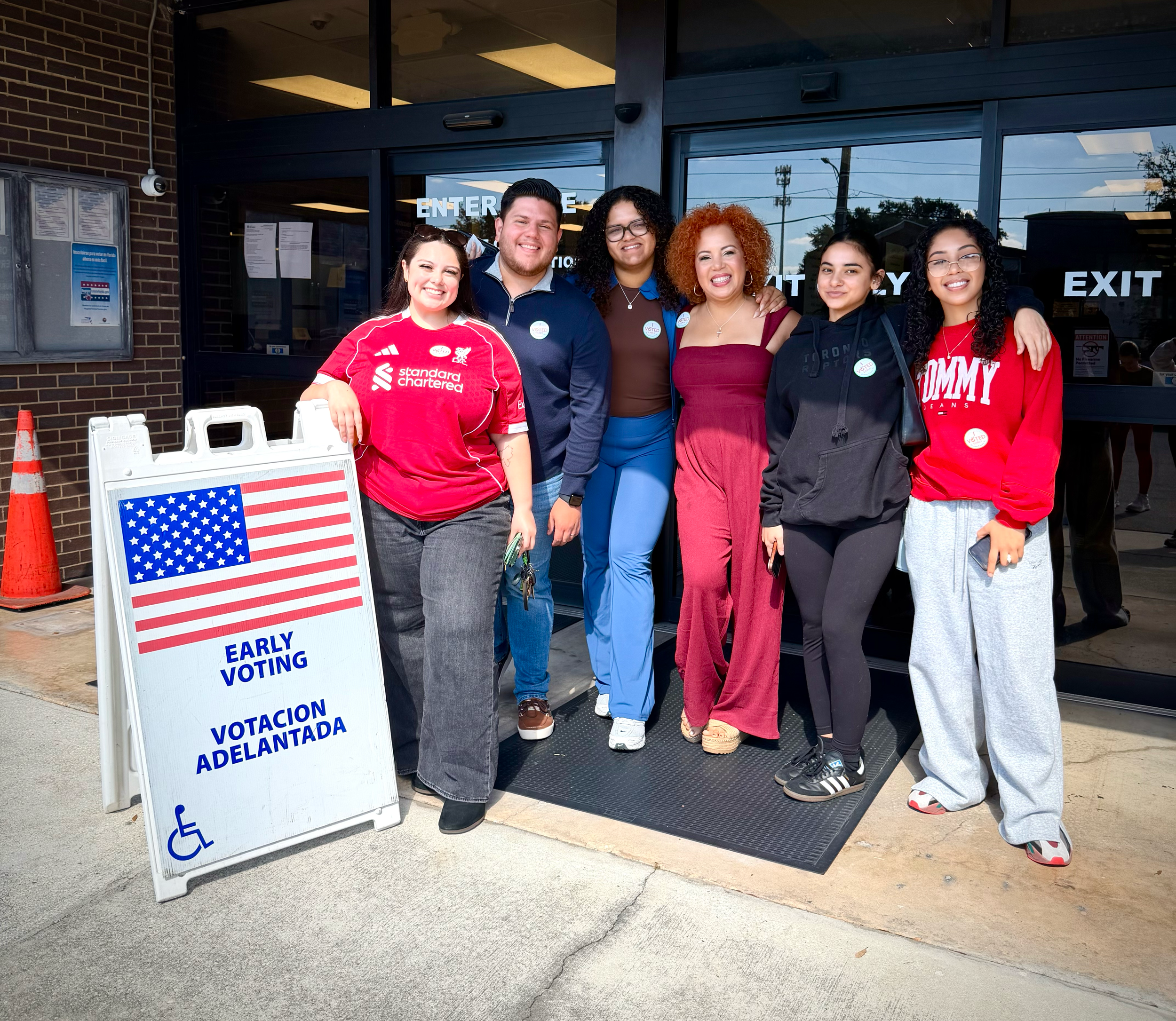 Six diverse people standing outside a building, smiling, with a sign that reads 'Early Voting' and 'Votacion Adelantada', indicating they are participating in early voting. The group includes both men and women, some wearing casual and others wearing