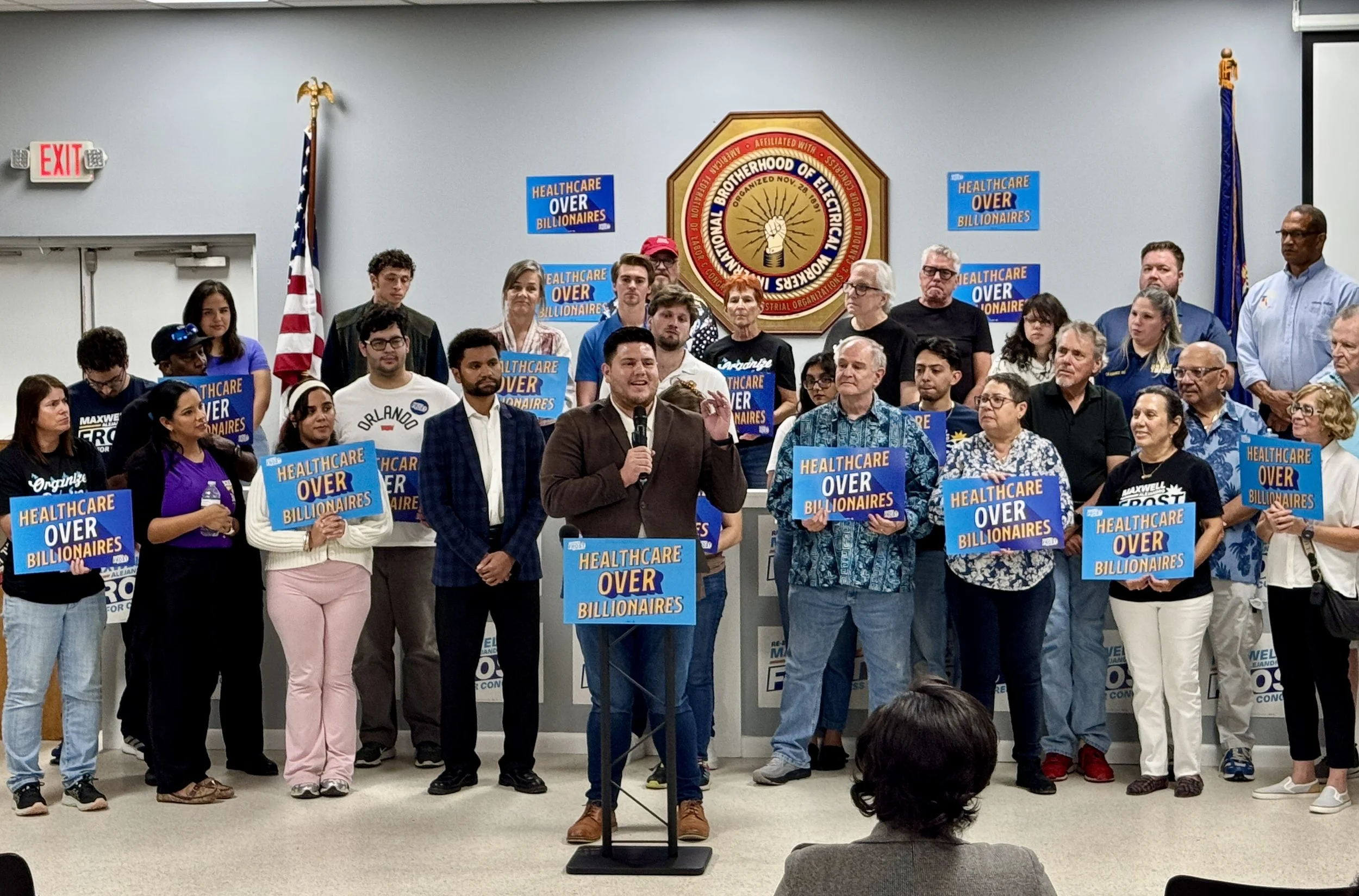 A group of people at a political rally with signs reading 'Healthcare Over Billionaires,' gathered in a room with a government emblem on the wall. A man in a brown jacket speaking at a podium, with a woman in a purple shirt holding a water bottle nearby.