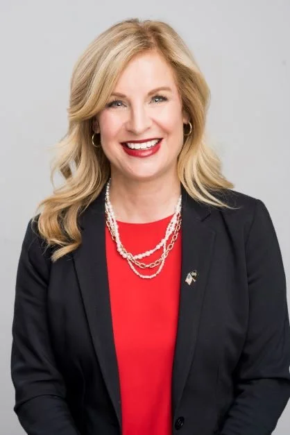 A woman with blonde hair wearing a black blazer, red top, and pearl necklaces, smiling at the camera.