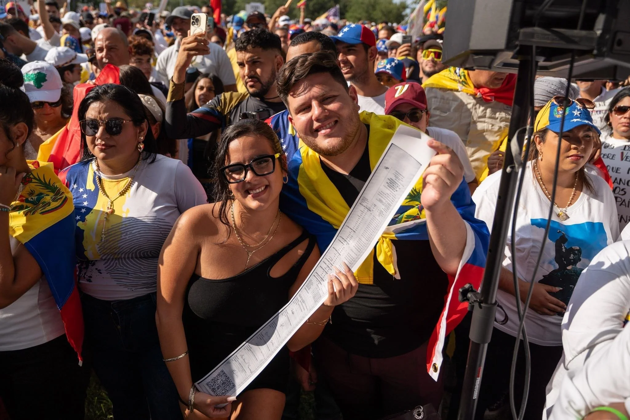 People at a crowded outdoor event, some wearing Venezuelan flag-themed clothing and accessories, holding a long printed paper, with one smiling person in the foreground.