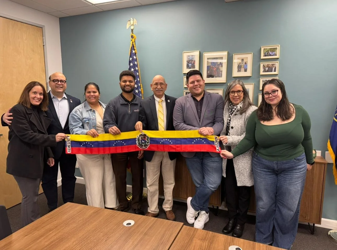 Group of nine diverse people standing in a meeting room holding a small flag with a coat of arms and red, blue, and yellow stripes, with an American flag and framed photographs in the background.