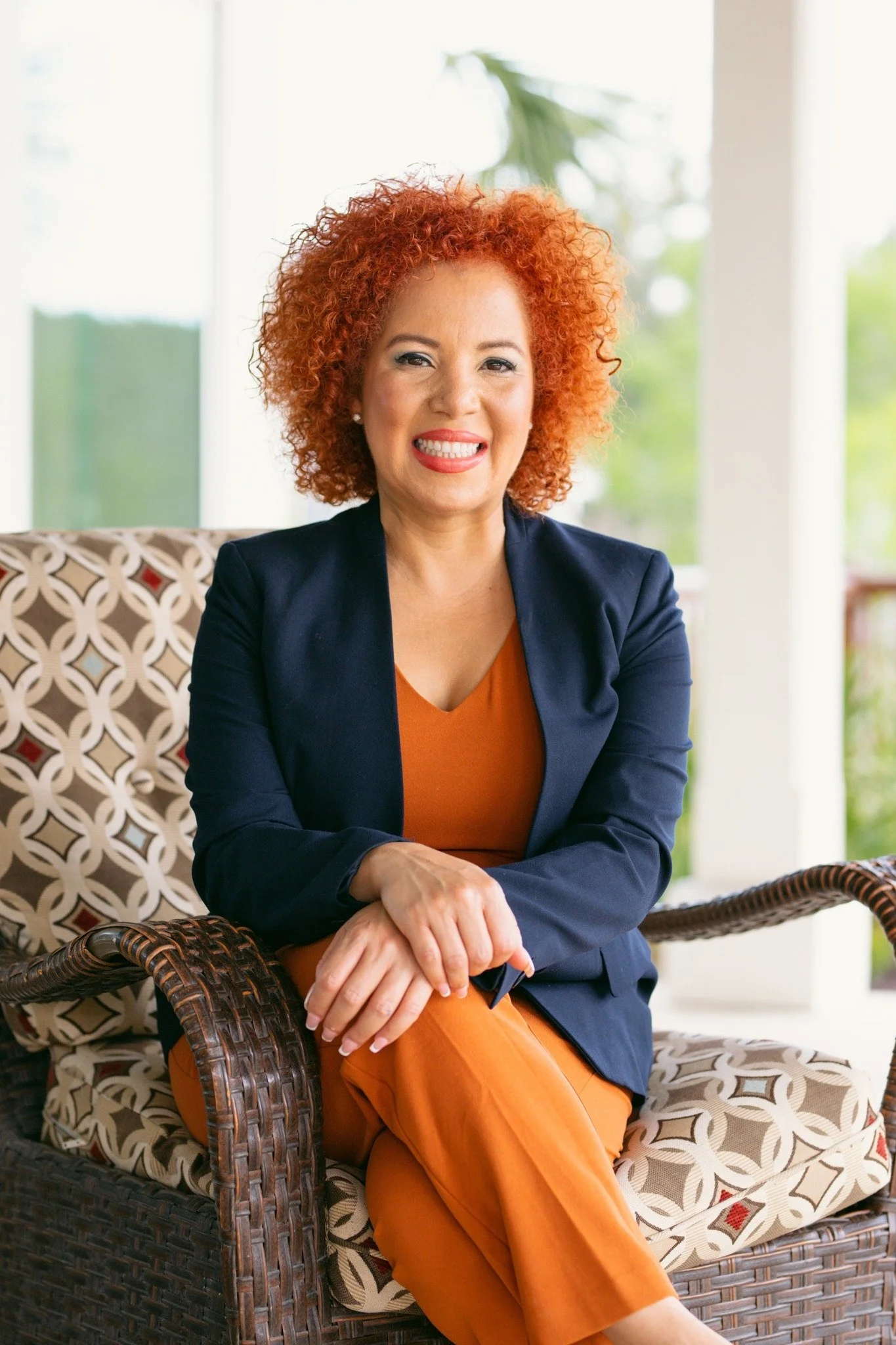 A woman with curly red hair sitting on a wicker chair with patterned cushions, smiling, wearing a navy blazer and orange outfit, in a bright room with large windows and greenery outside.