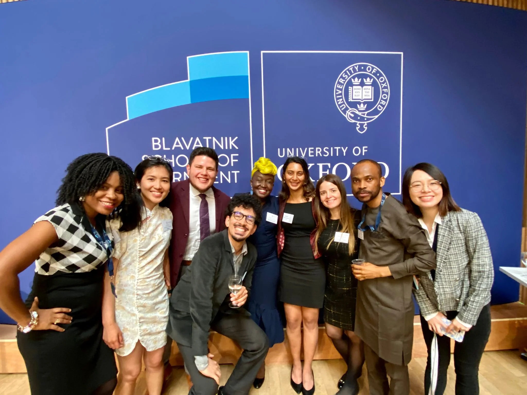 A group of nine diverse people posing for a photo in front of a blue backdrop with the logos of Blavatnik School of Government and University of Oxford. They are smiling and appear to be at a formal or professional event.