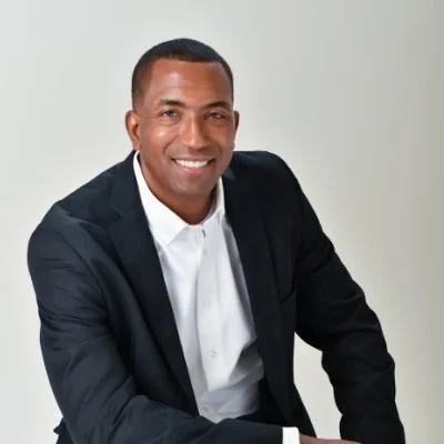 A smiling man in a dark suit and white shirt, seated against a plain light gray background.
