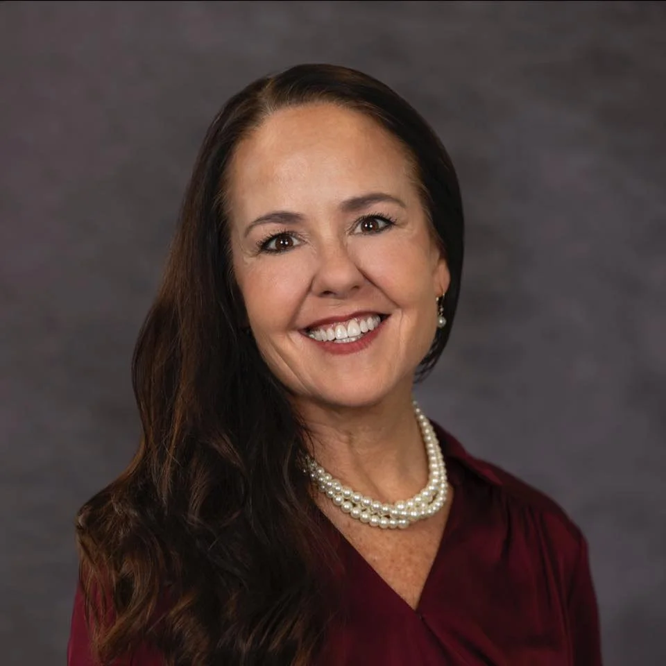 A woman smiling, with long dark hair, wearing pearl earrings, a pearl necklace, and a burgundy top, standing against a gray background.