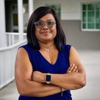 Woman with black hair wearing glasses, a blue sleeveless top, and a cross necklace, standing outdoors with arms crossed.