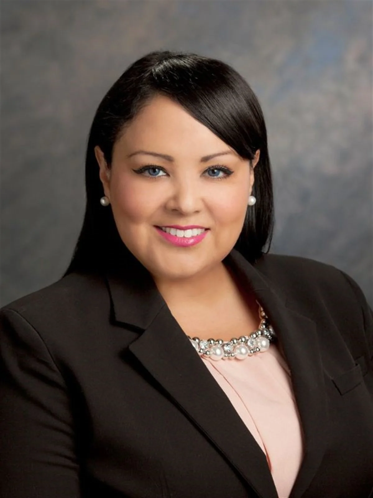 Professional portrait of a woman with black hair, wearing a black blazer, pearl earrings, a pearl necklace, and pink lipstick, smiling against a gray background.