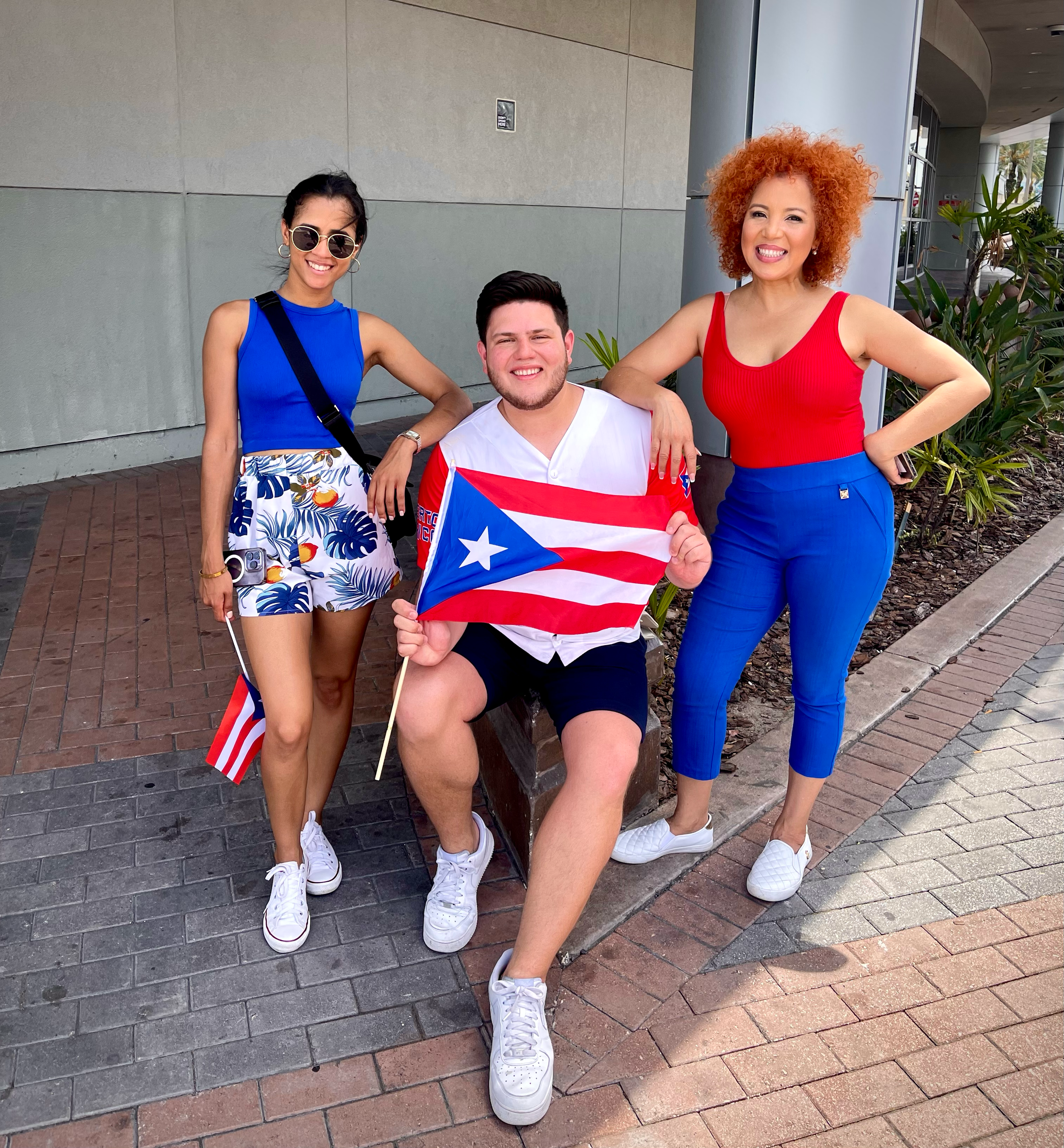 Three people celebrating Puerto Rican independence outdoors. One man sitting on a bench holding a Puerto Rican flag, and two women standing on either side, one with a small Puerto Rican flag, all dressed in red, white, blue, and casual summer clothin
