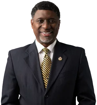 A smiling man in a dark suit, white shirt, and striped yellow tie, standing against a black background.