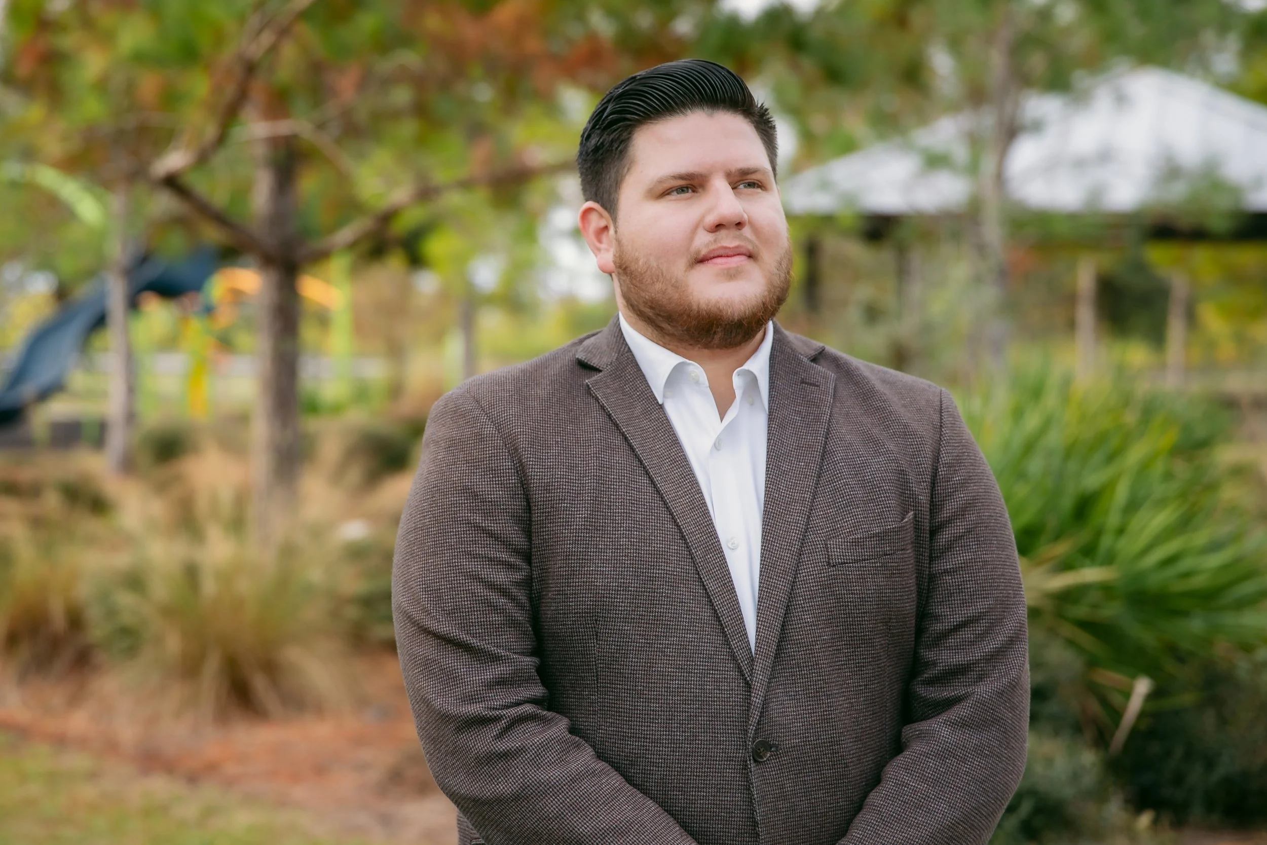 A man in a brown suit jacket and white shirt standing outdoors with trees and greenery in the background.