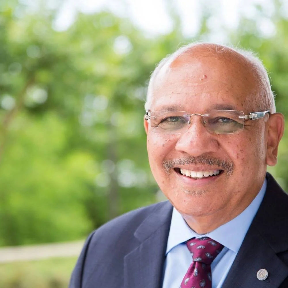 A smiling man wearing glasses, a blue shirt, a maroon tie, and a dark suit, with a blurred green outdoor background.