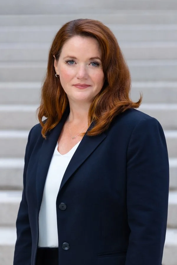 A woman with shoulder-length red hair, wearing a navy blazer over a white top, standing outdoors in front of steps.