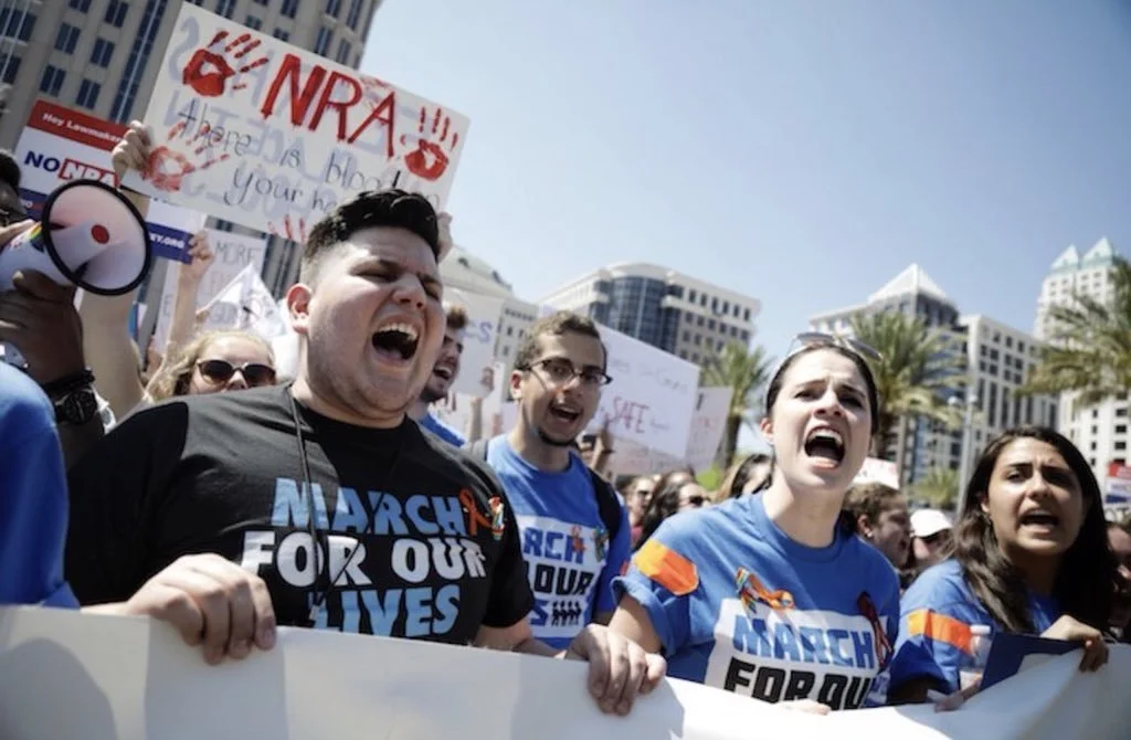A group of people protesting outdoors, holding signs and shouting. One sign reads 'NRA' with red handprints. Some are wearing blue shirts that say 'March For Our Lives'. There are tall buildings and palm trees in the background on a sunny day.
