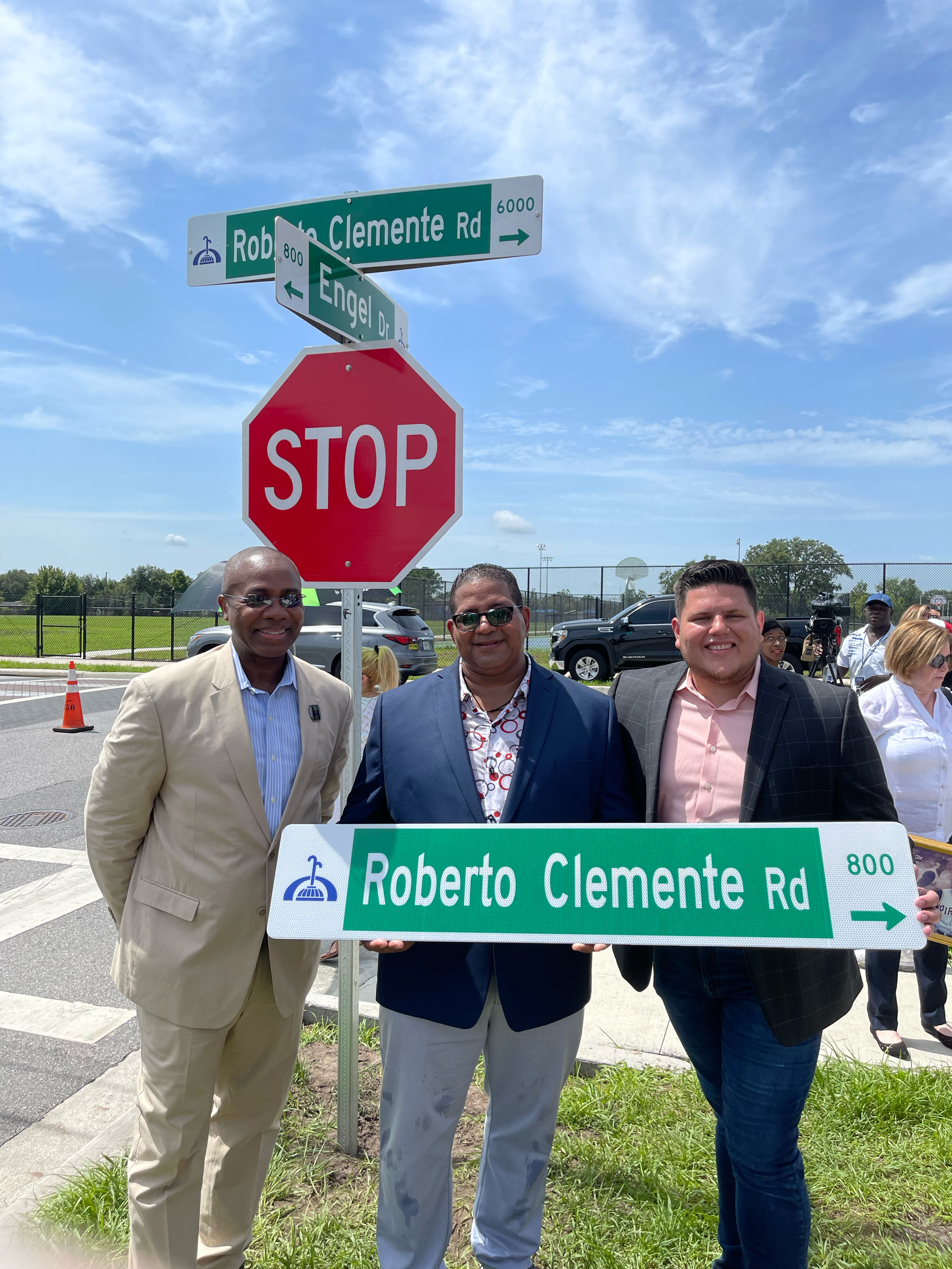 Three men in suits holding a street sign that reads 'Roberto Clemente Rd' at an outdoor event during the day, with stop sign and street signs visible in the background under a blue sky.