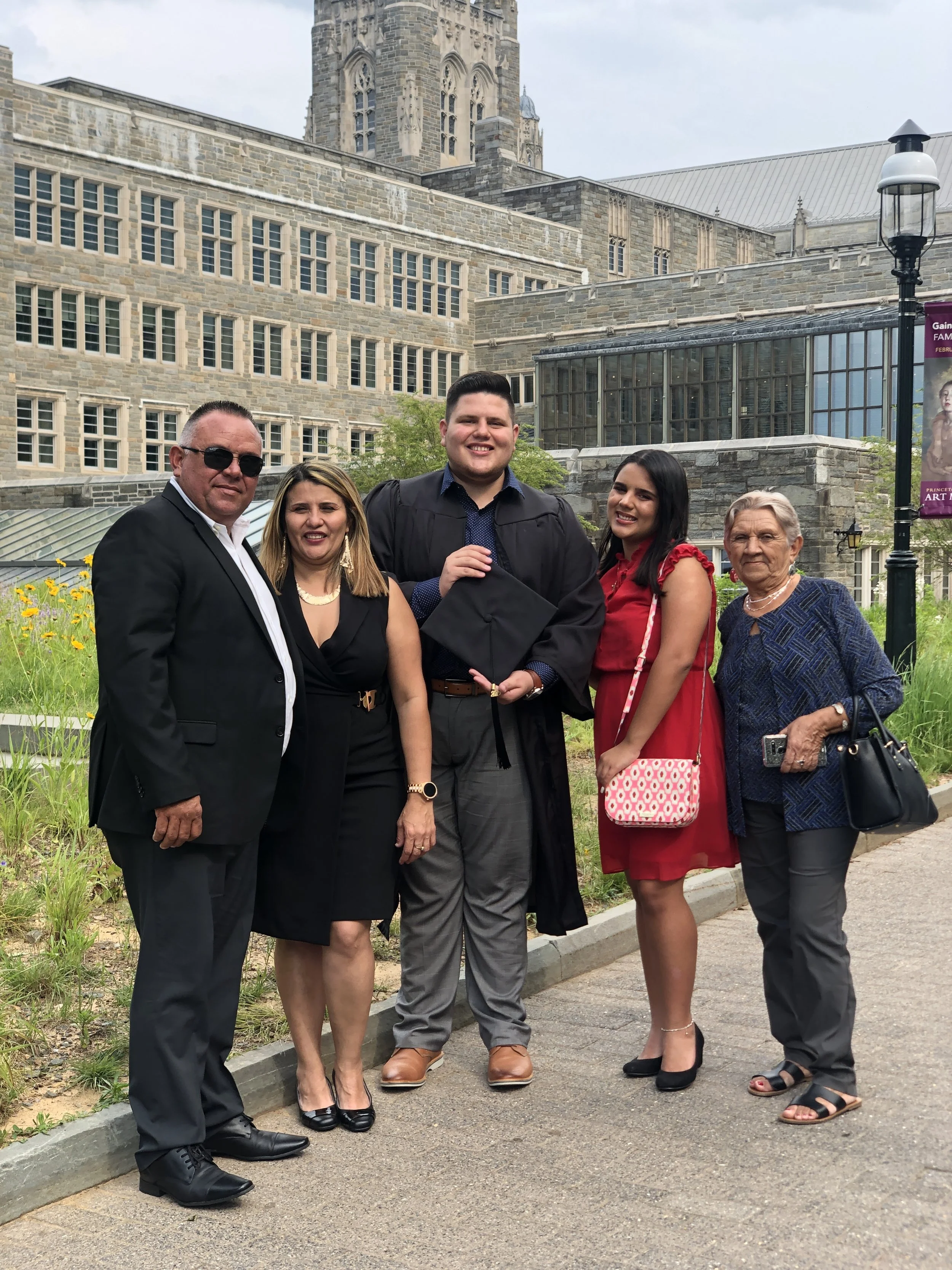 Group of five people celebrating a graduation outdoors in front of a historic building with a tower, smiling for the photo. The young man in the center is wearing a cap and gown.