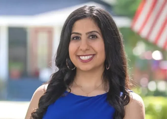 Young woman with dark hair and a bright smile, wearing a blue top, outdoors with blurred colorful background.