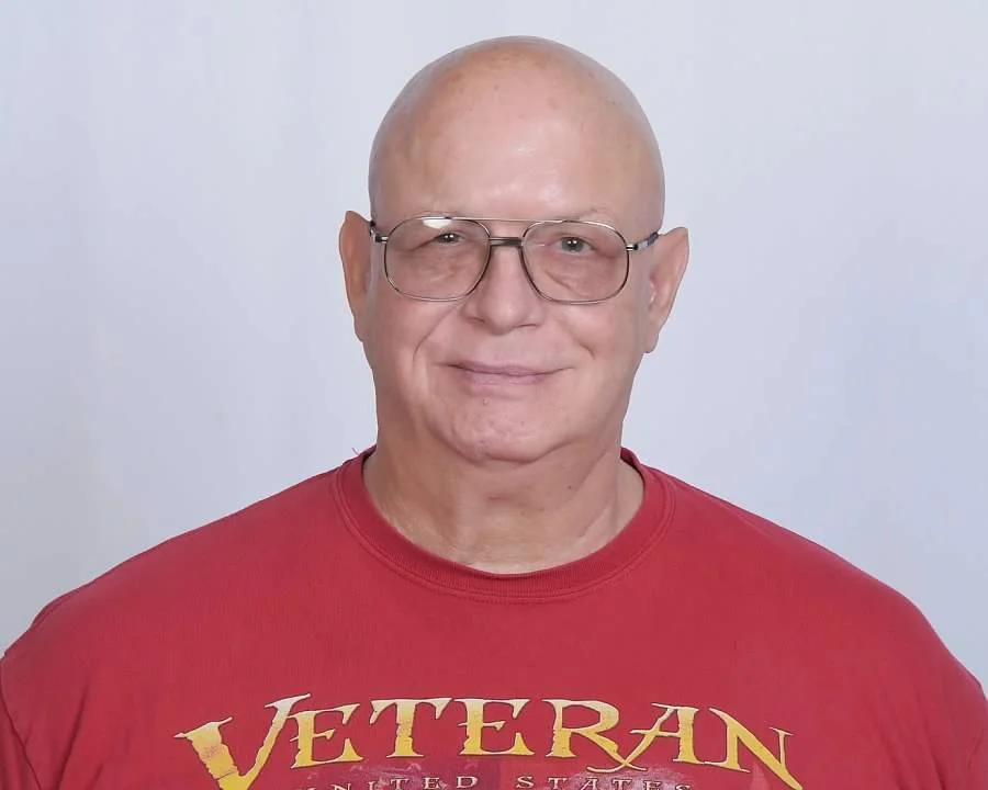 A smiling bald man wearing glasses and a red T-shirt with the word "VETERAN" printed on it, standing against a plain white background.
