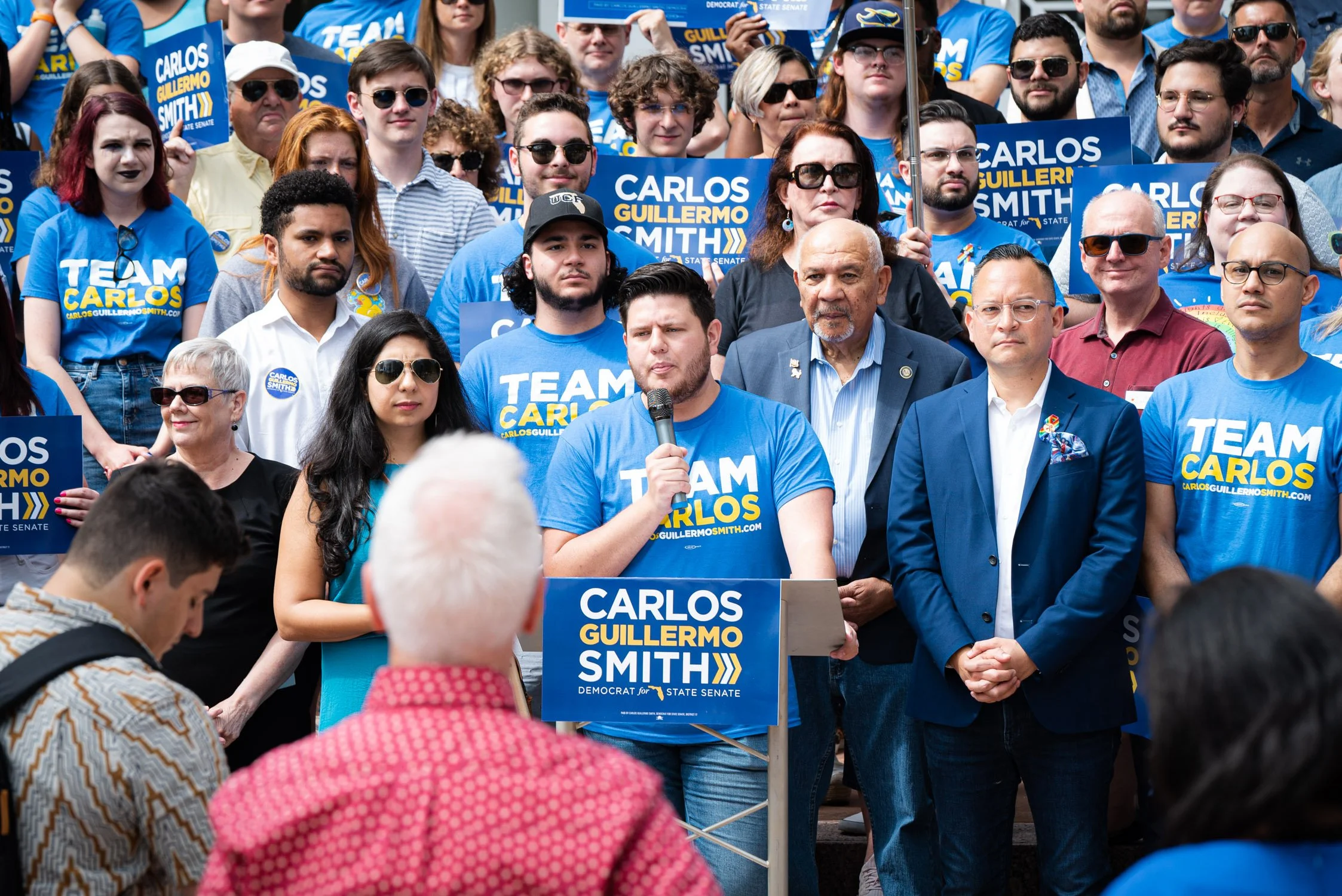 A group of people at a political rally, many wearing blue shirts with 'TEAM CARLOS' and 'CARLOS GUILLERMO SMITH' on them. A young man is speaking into a microphone at a podium with the same campaign signage. The speaker and audience members appear en