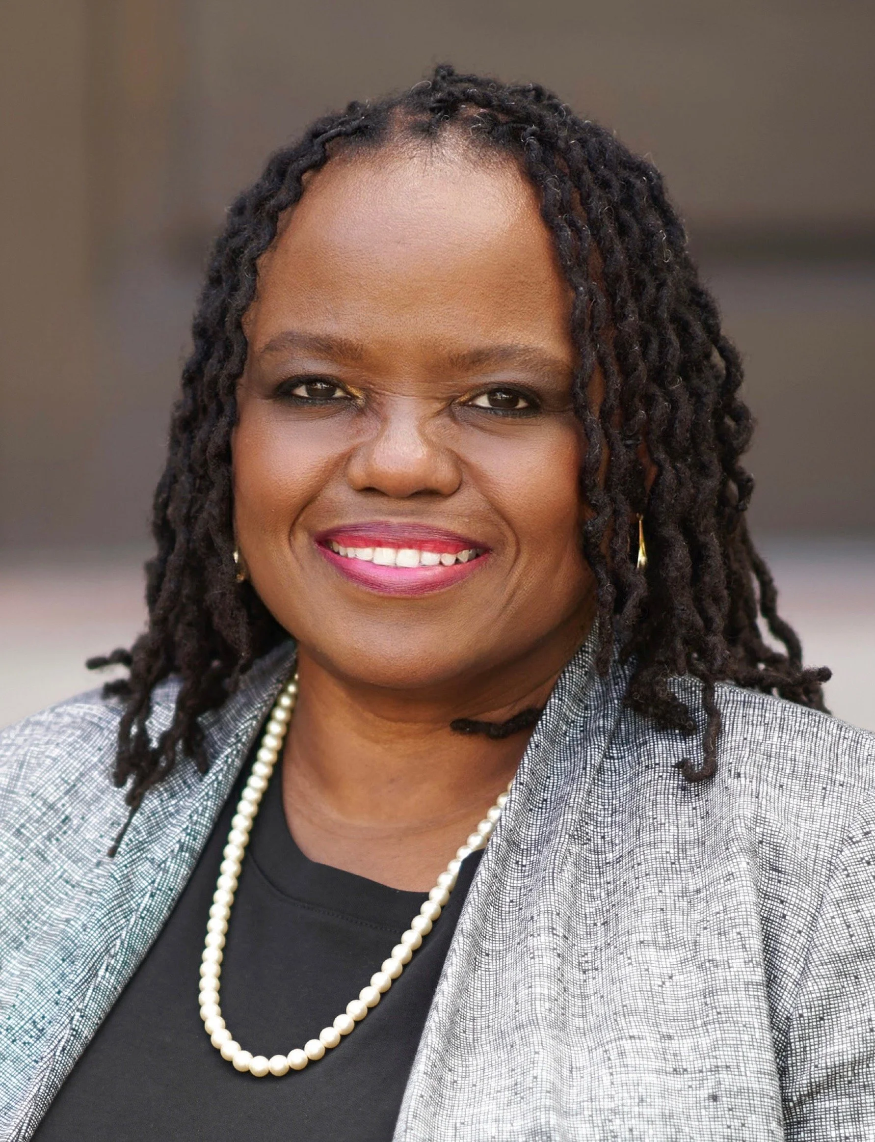 Close-up photograph of an African American woman with shoulder-length curly hair, wearing a gray blazer, a black top, a pearl necklace, and gold earrings, smiling at the camera.
